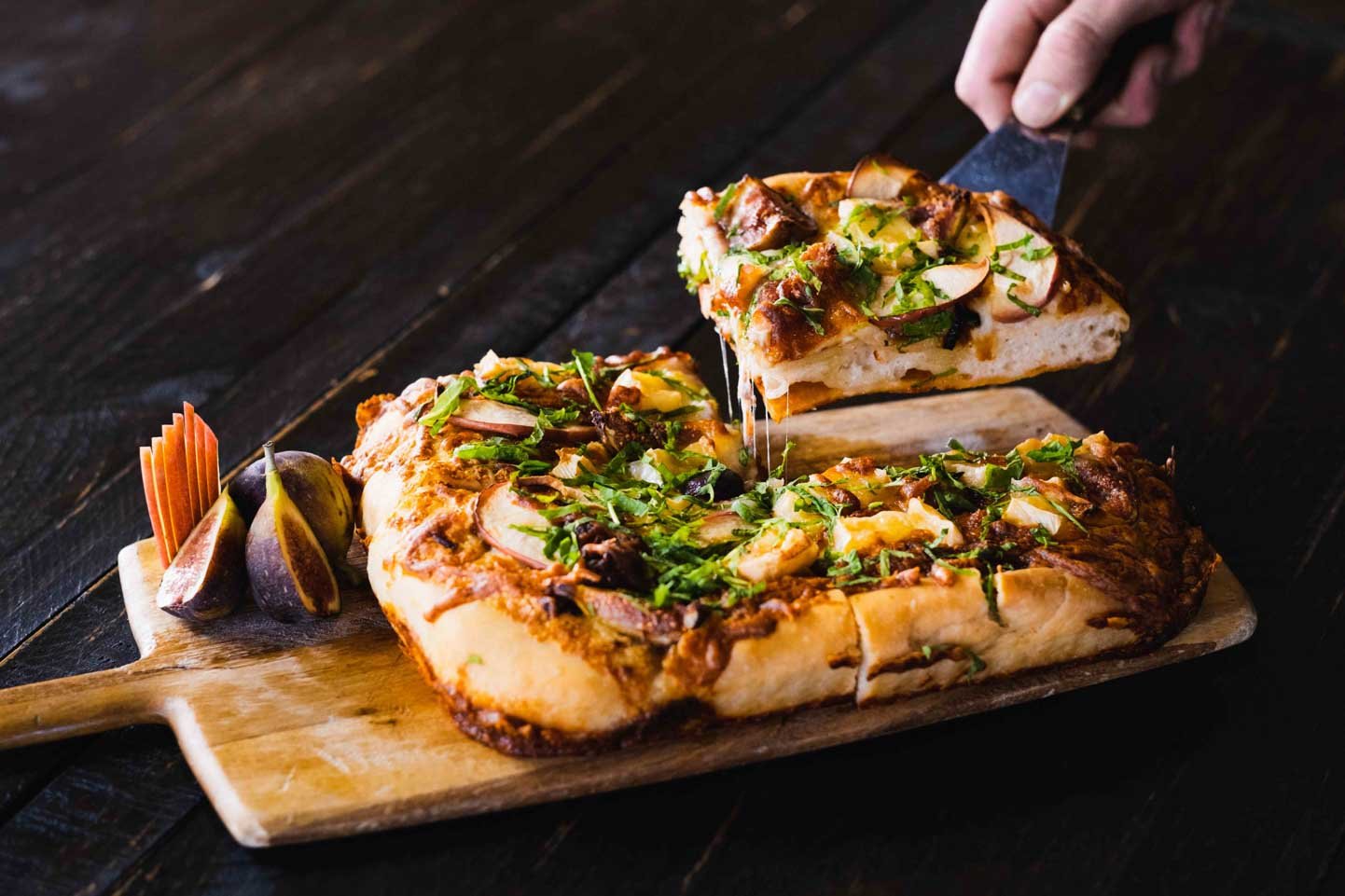 A person serving a slice of pizza with chopped herbs and vegetables on a wooden board, with a side of figs.
