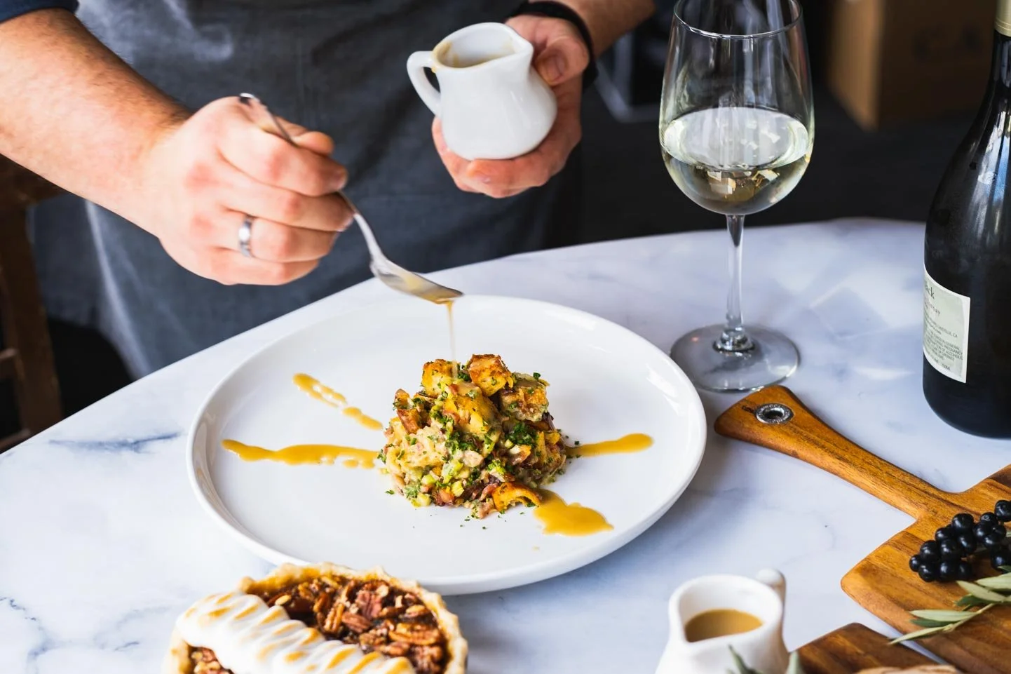 Person drizzling sauce over a plated dish of potato salad on a white marble table, with a glass of white wine, a small pitcher of sauce, and a wooden board with grapes and a bottle of wine.
