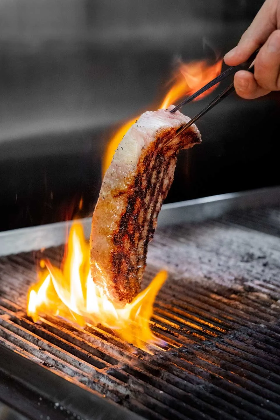 A person using tongs to hold a piece of bacon over an open flame on a grill, with flames and smoke.