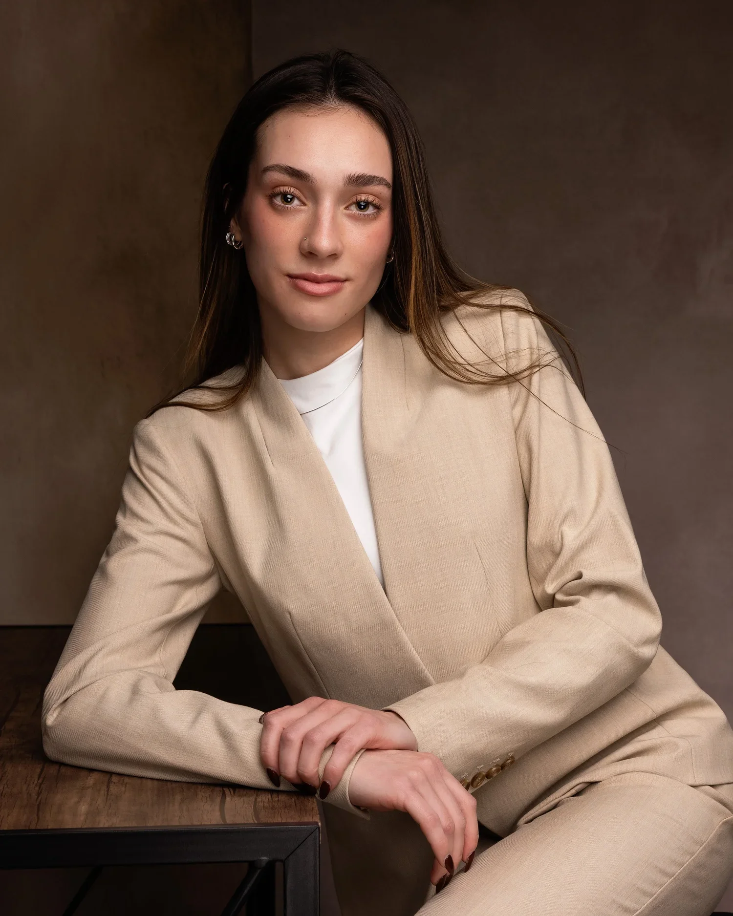 A young woman with long brown hair, wearing a beige suit jacket and white shirt, sitting with her arm resting on a table, looking at the camera against a brown background.