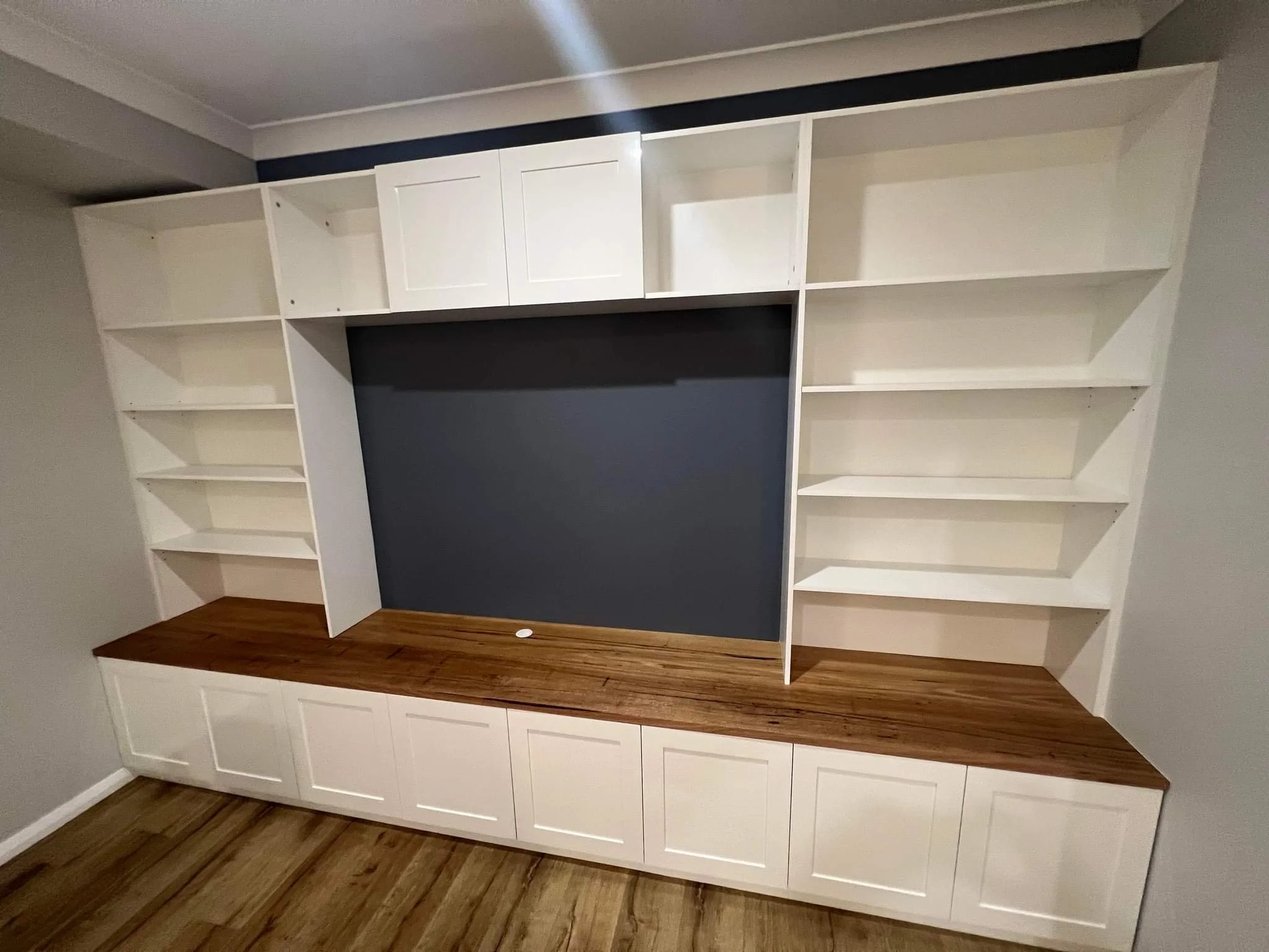 Built-in white shelving unit with upper cabinets, open shelves, and a wooden countertop, installed against a dark accent wall and adjacent to a light wood floor.
