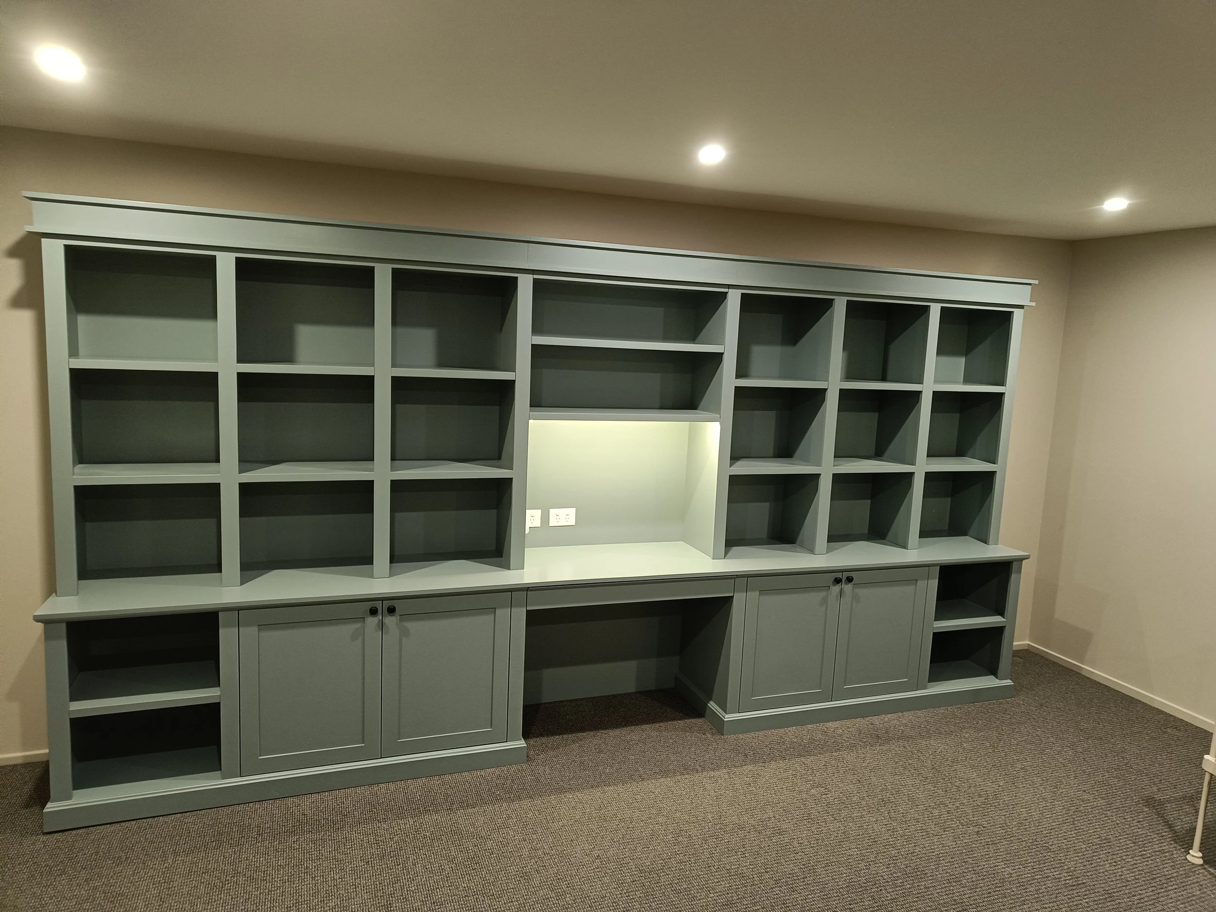 Custom-built green bookshelf and desk unit in a room with recessed lighting and carpeted floor.