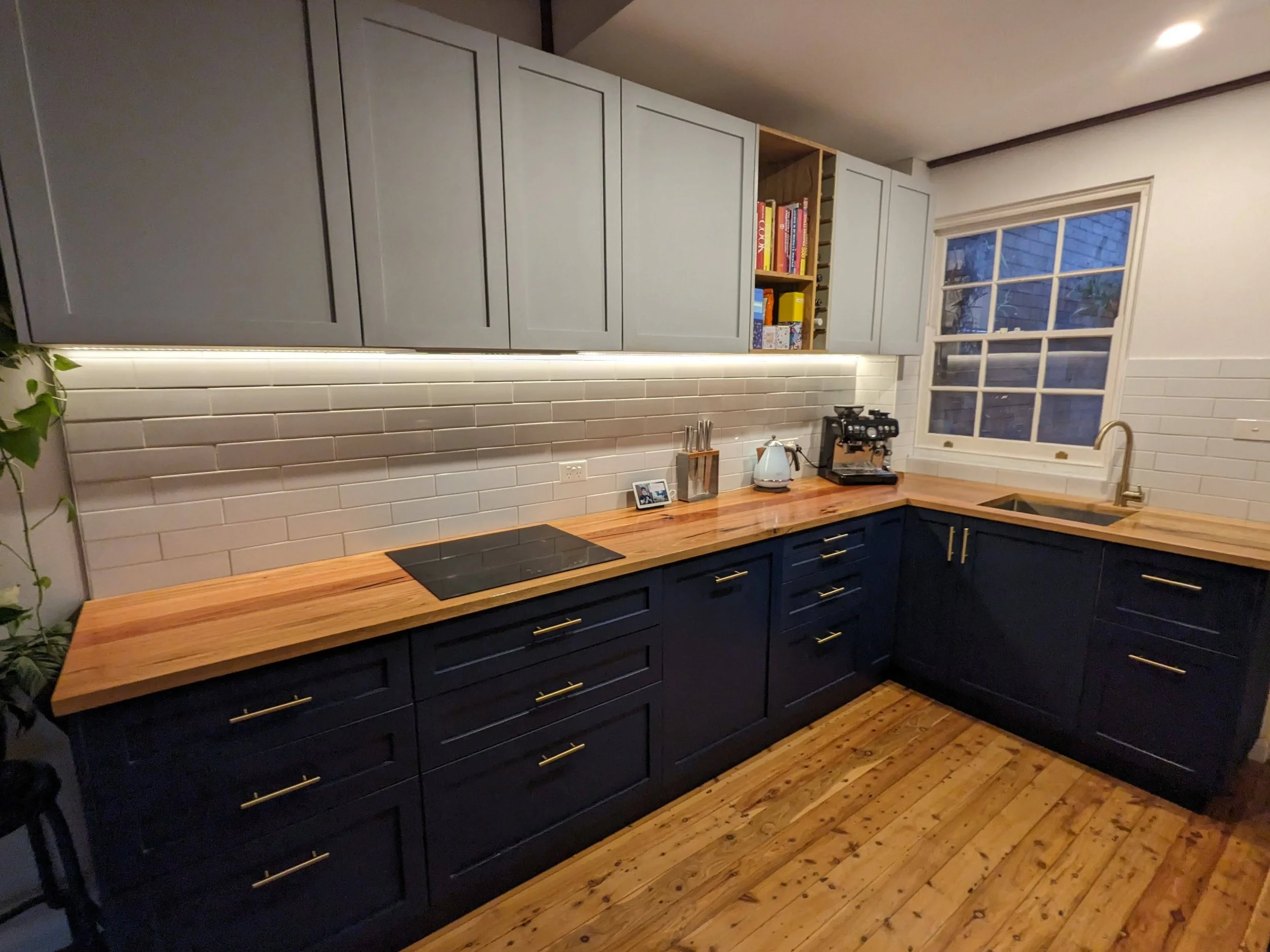 Modern kitchen with wooden countertops, dark blue cabinets, white subway tile backsplash, and integrated appliances. Features include an induction cooktop, coffee machine, and kettle. Upper cabinets have under-cabinet lighting. Window with a view.