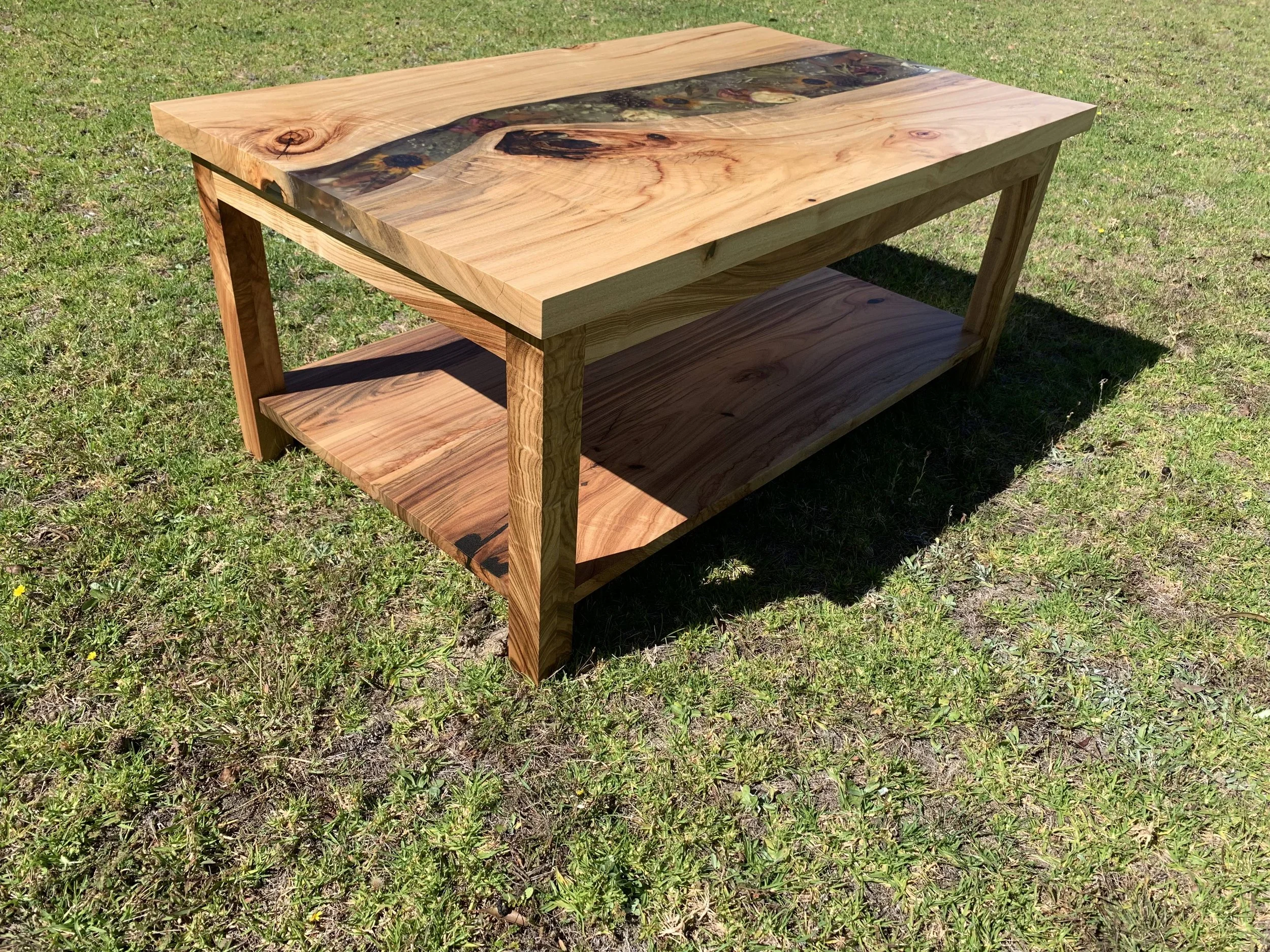 Wooden coffee table with natural finish and epoxy resin river on the top, set on grassy ground.
