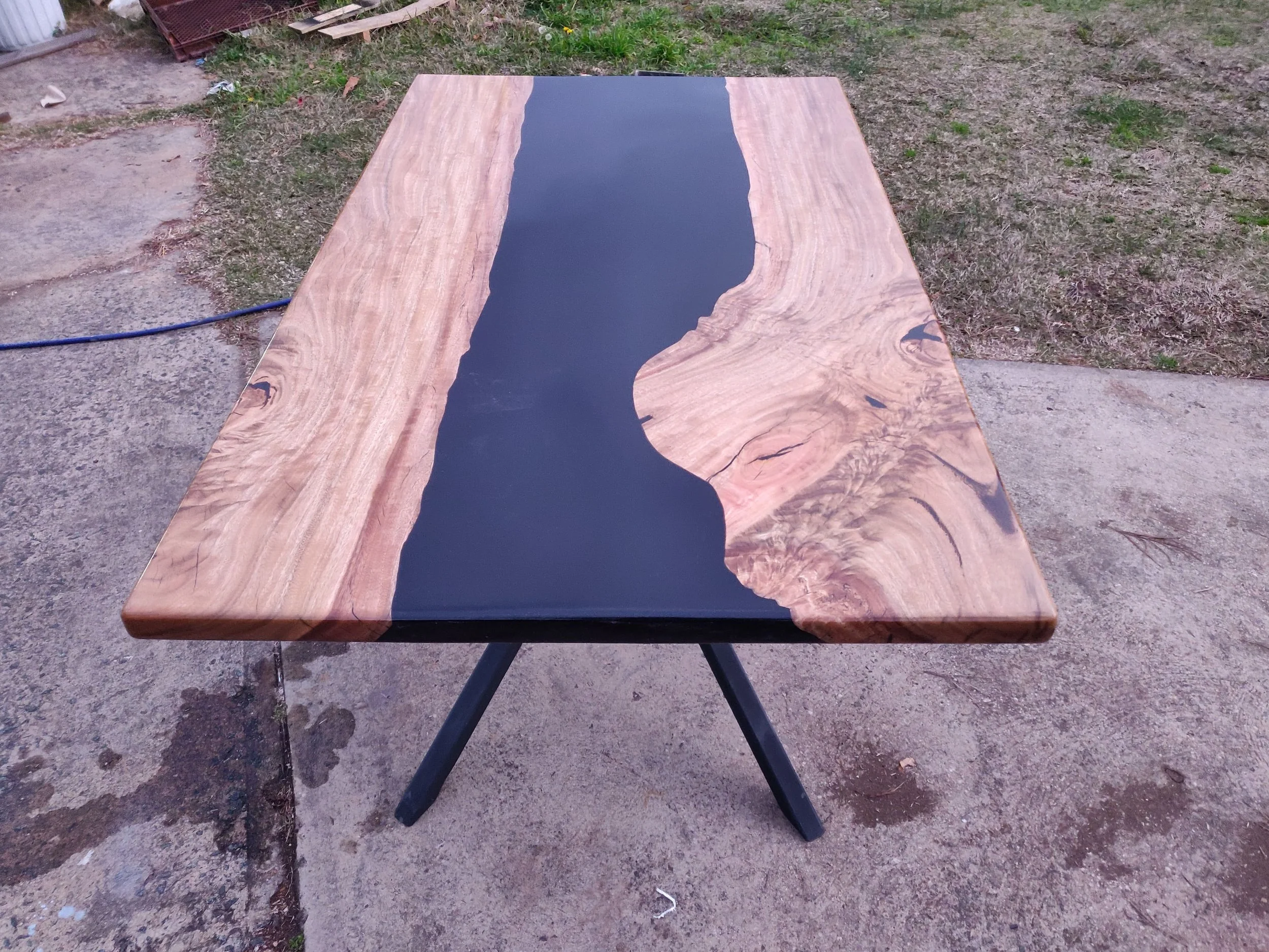 Wood and epoxy resin table with black center and natural wood edges on a angled metal base, placed on outdoor concrete surface.