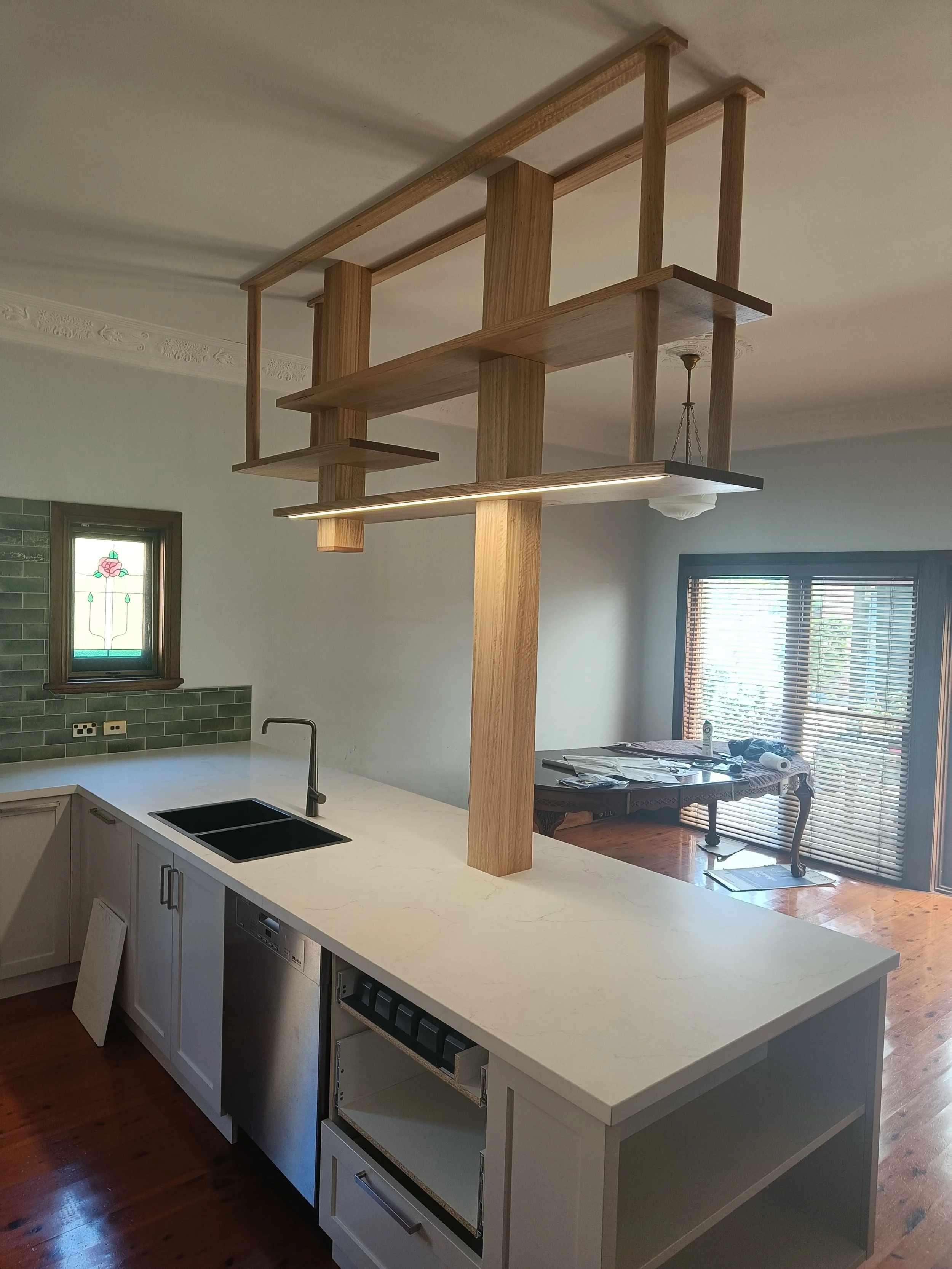 Modern kitchen with large island, featuring a light wooden frame structure above, a black faucet, a white countertop, built-in dishwasher, and green tiled backsplash.
