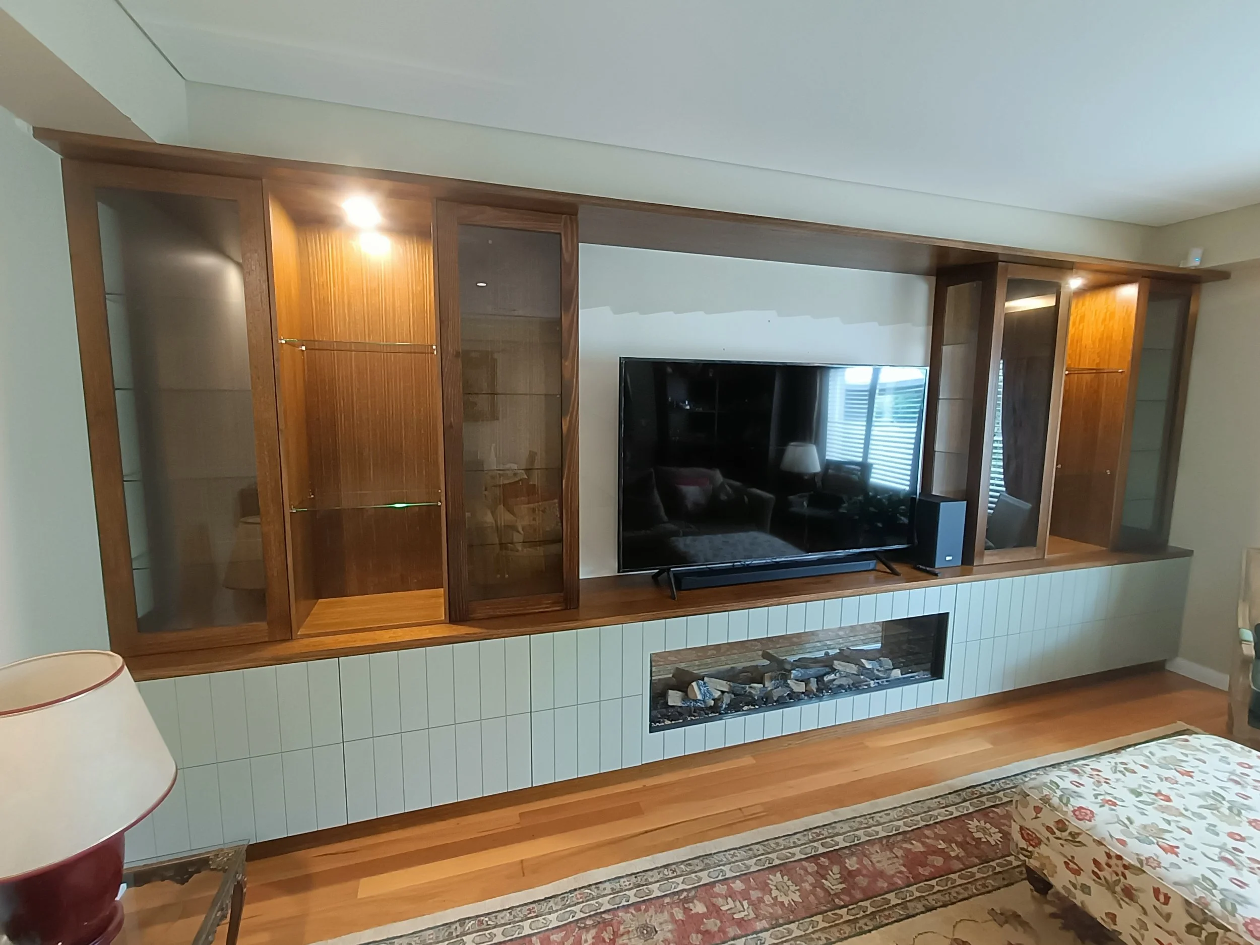 Modern living room showcasing a built-in wooden entertainment center with glass cabinets, featuring a large flat-screen TV and an electric fireplace below, with decorative items on side tables and an upholstered ottoman in the foreground.