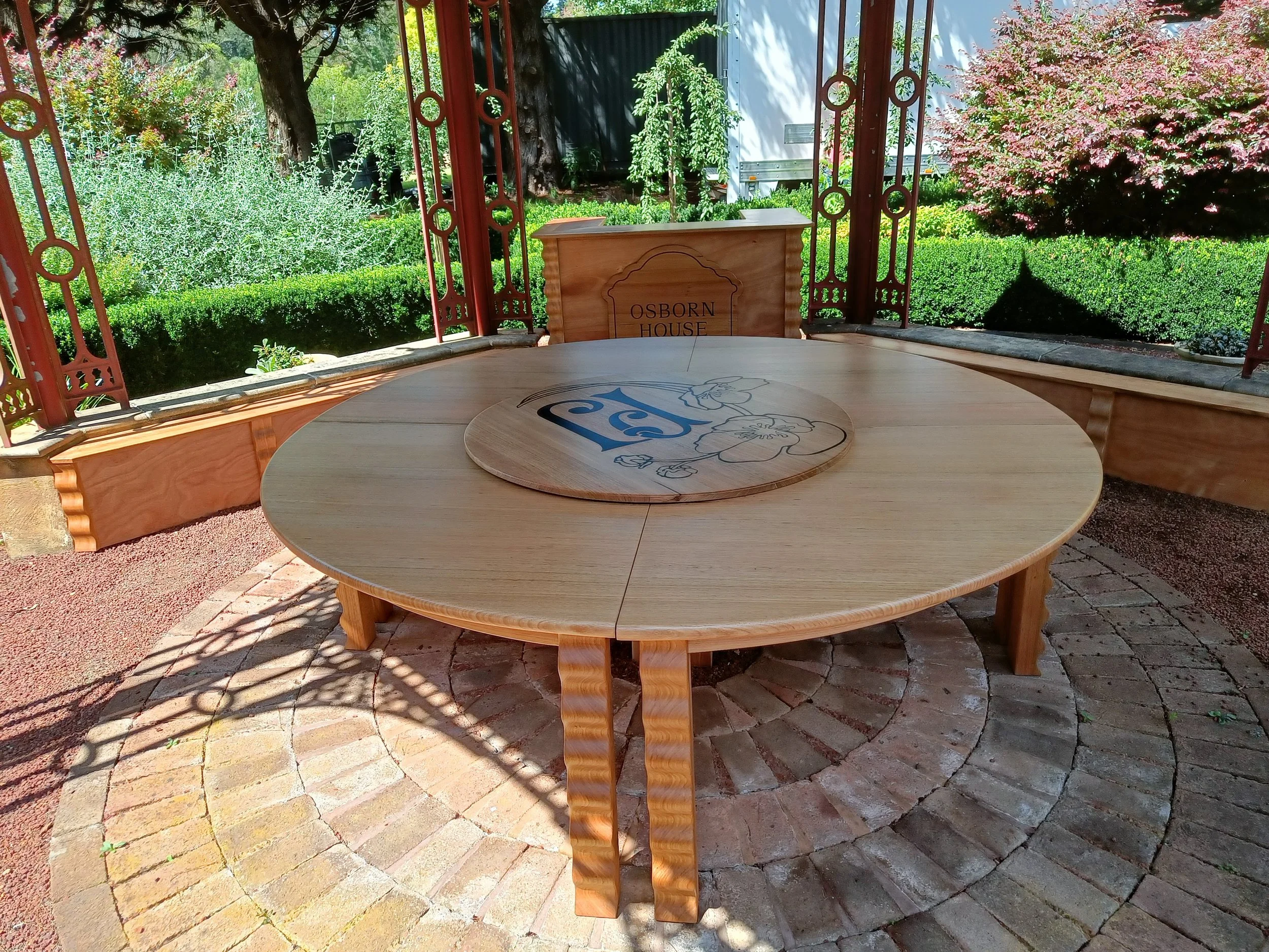 A round wooden table with a decorative inlaid design is placed on a brick patio within a gazebo. The backdrop includes a manicured garden with trees, hedges, and flowering bushes. The gazebo has ornate metal detailing and a sign that reads "Osbourn H