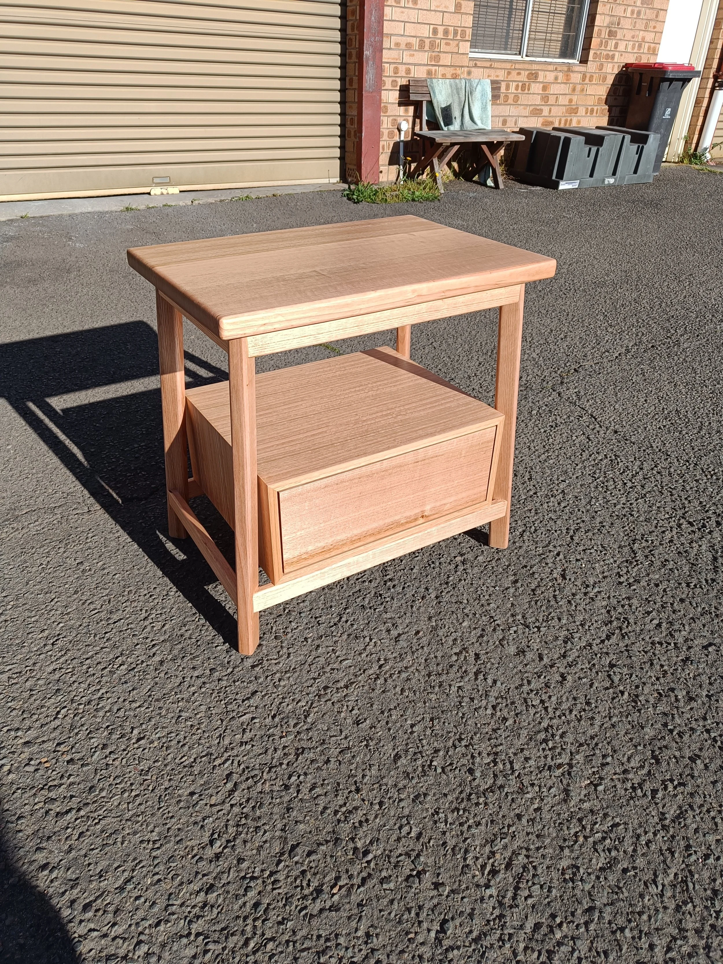 Wooden side table with a drawer placed outdoors on pavement; background includes a brick building, metal garage door, and utility bins.