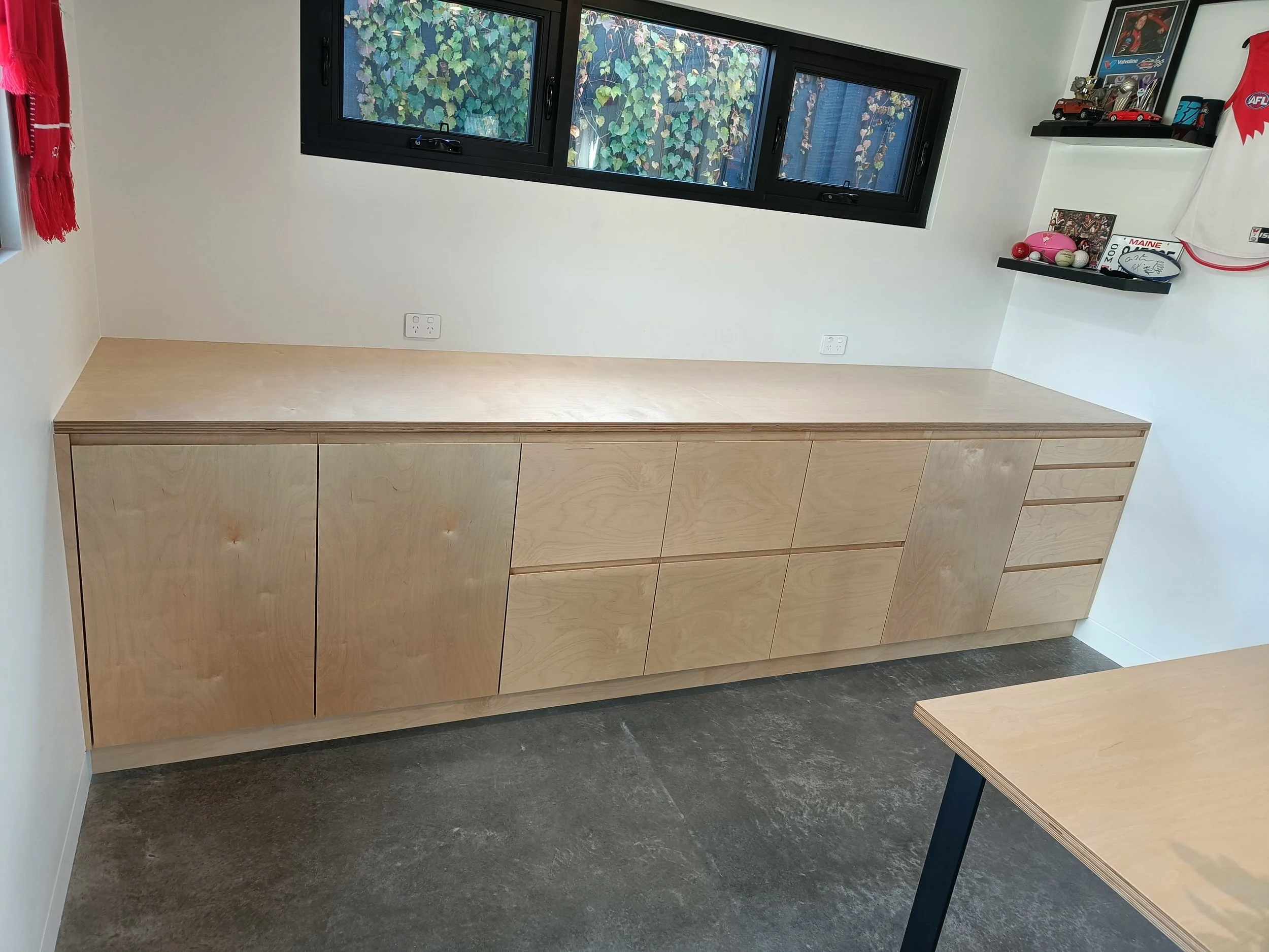 Minimalist wooden cabinetry with drawers and cupboards under a black-framed window next to a wall with shelves and decor items.