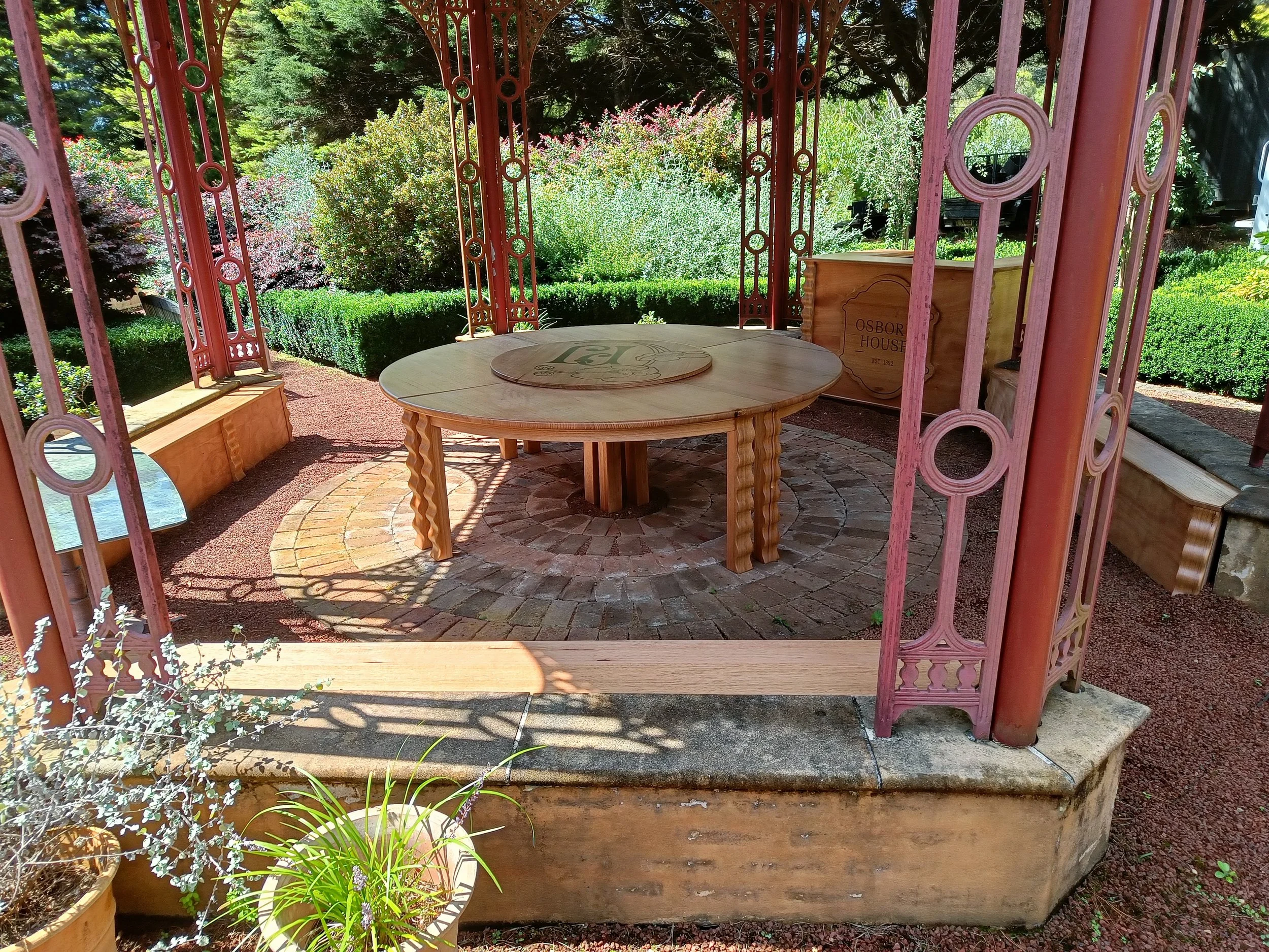 A gazebo with a round wooden table and benches, surrounded by metal lattice structures. The floor is made of circular brick patterns and there's greenery outside the gazebo. Planters with plants are placed inside as well. Sunlight is casting shadows