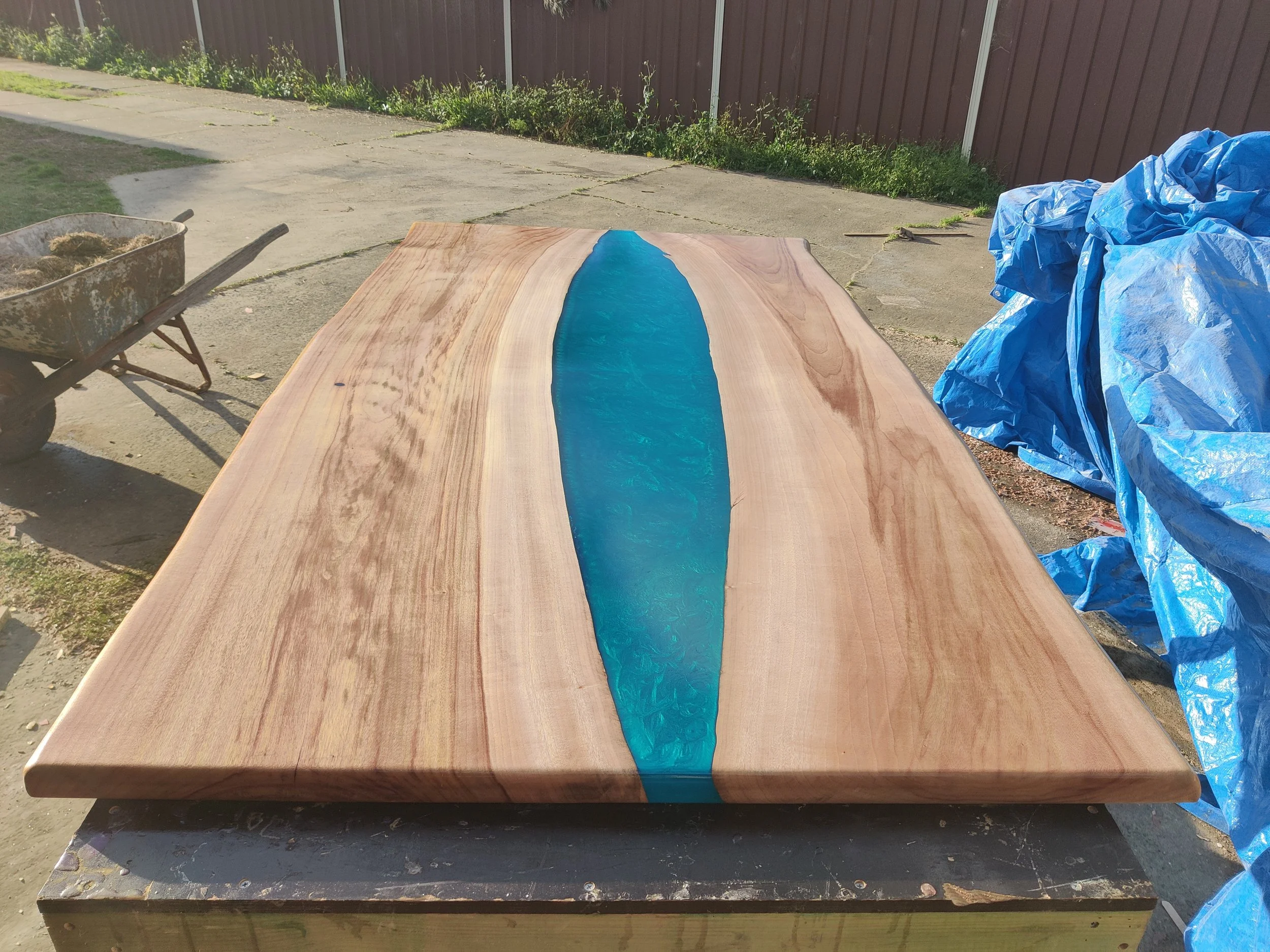Wooden tabletop with blue epoxy resin river design, set outdoors on a workbench, adjacent to a wheelbarrow and blue tarps.