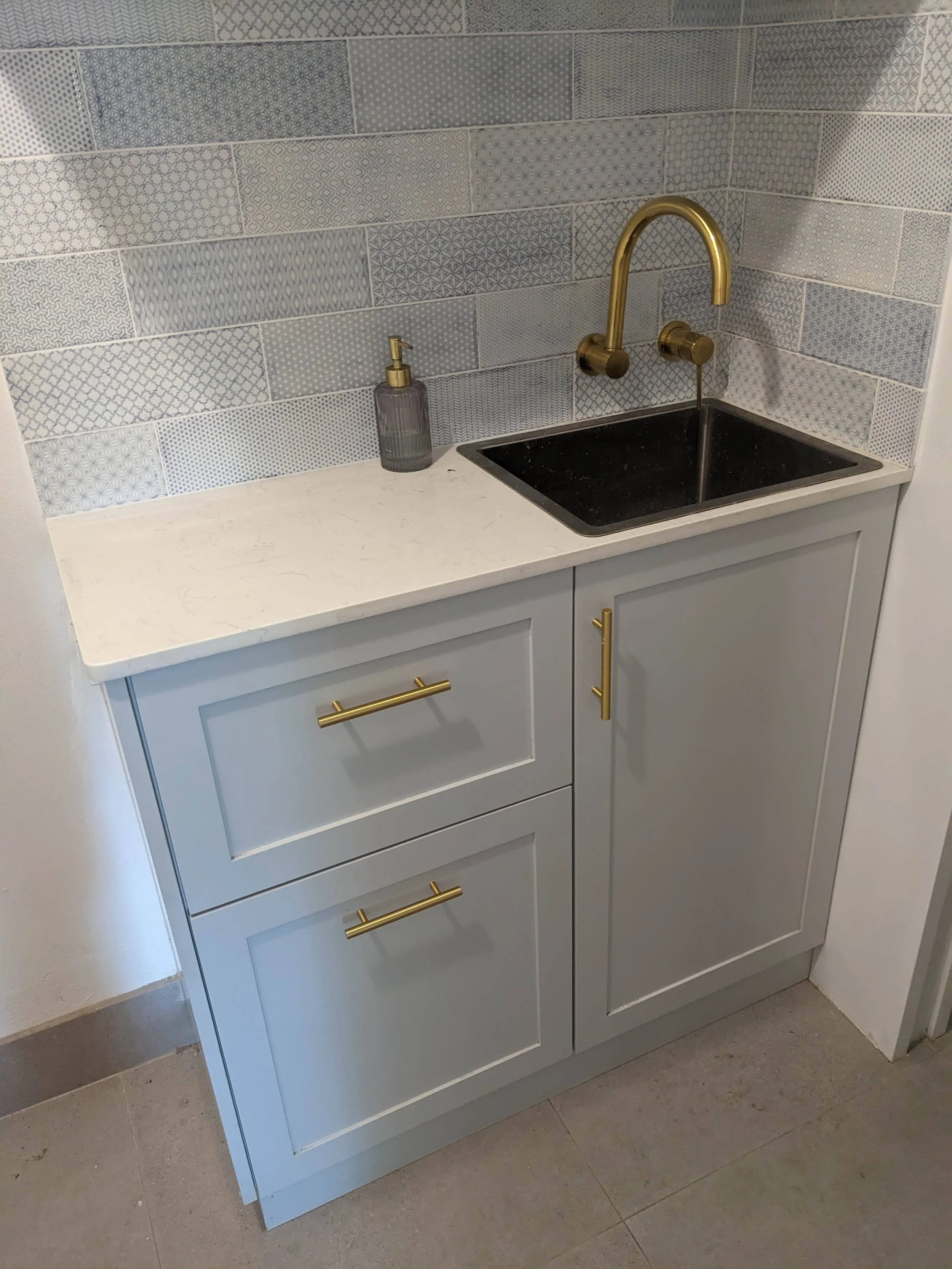 Bathroom vanity with blue patterned tile backsplash, a gray countertop, a black sink with a brass faucet, and gold hardware on blue cabinets.