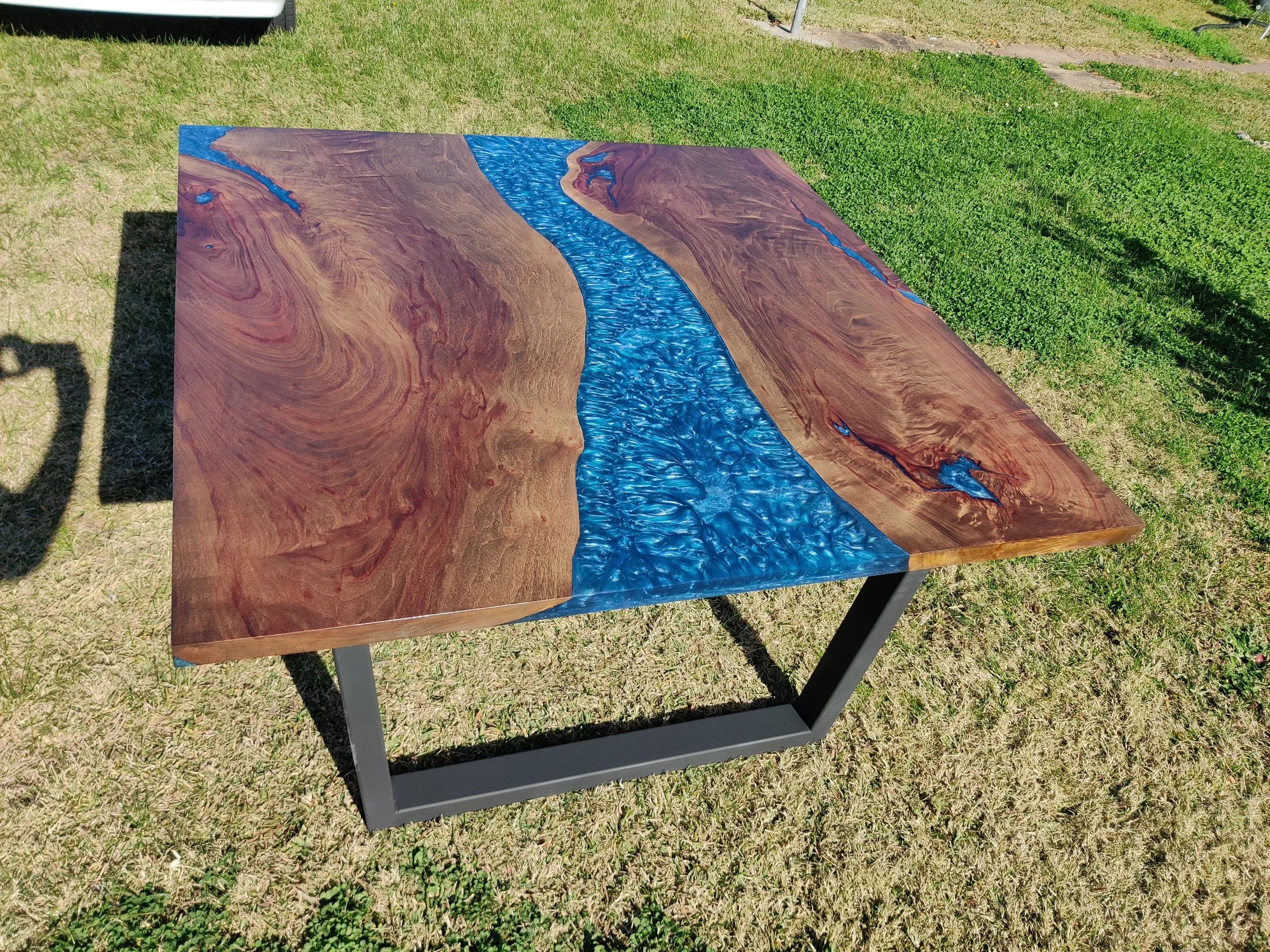 Wood and resin river table with a blue epoxy inlay on a metal base, placed on grass.