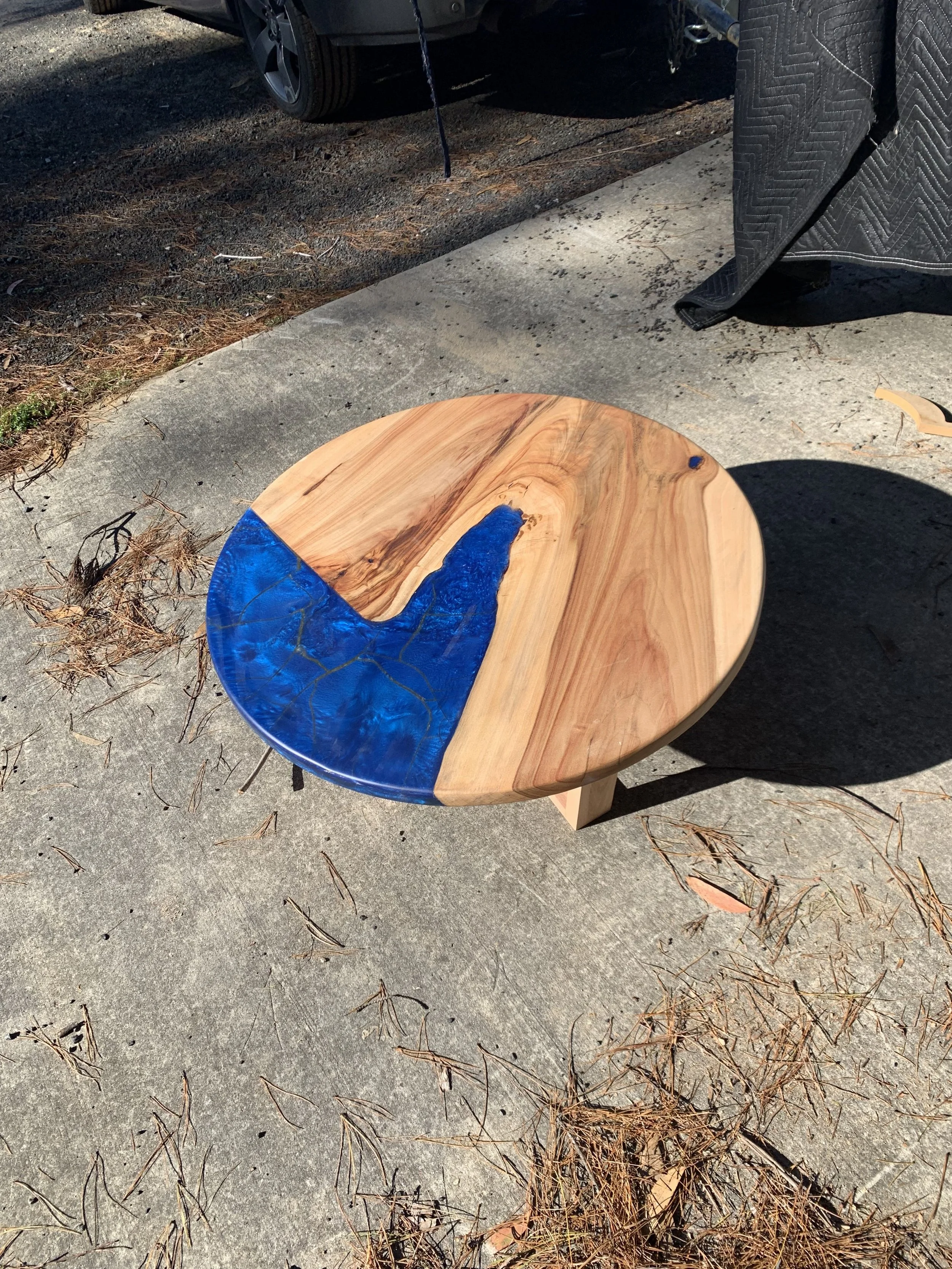 A round wooden table with a blue resin inlay on a concrete surface outdoors, with a vehicle and black cloth in the background.