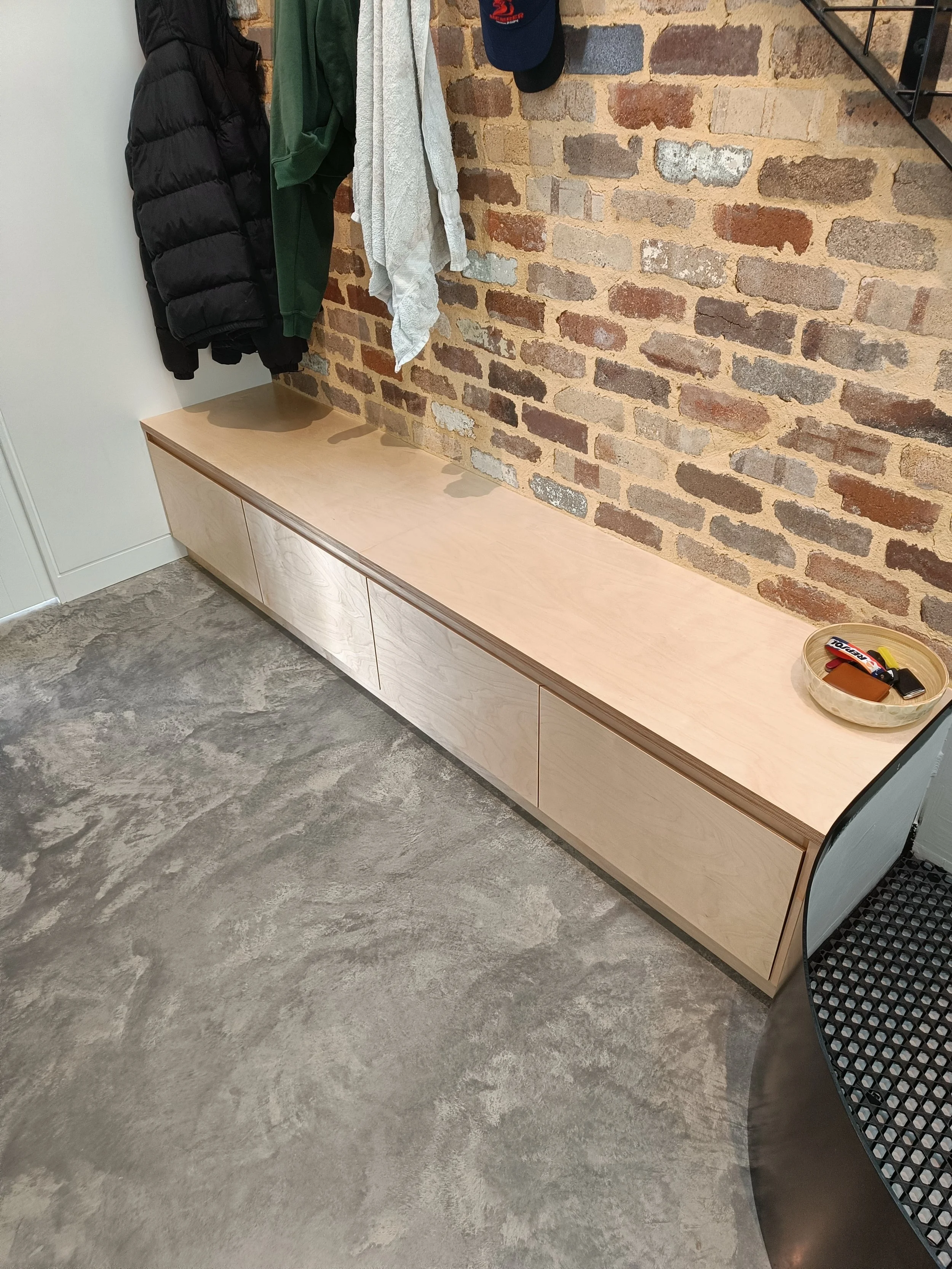 Wooden storage bench against brick wall, coats and hats hanging above, gray concrete floor, and a small dish with items on bench.