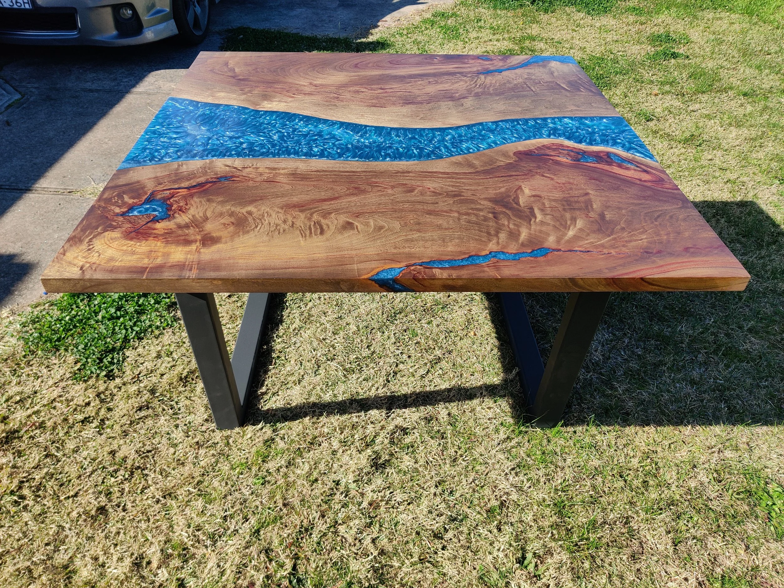 Wooden table with blue resin river design on metal legs, placed on grass.