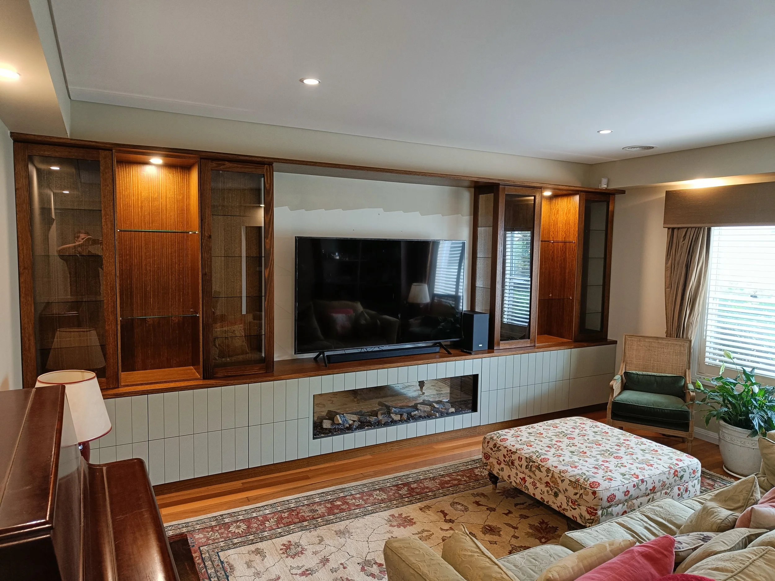 Living room with a wall-mounted TV, wooden shelves, and a decorative ottoman. A sofa, armchair, and piano are also visible. The room has wooden flooring, a patterned rug, and a plant by the window.