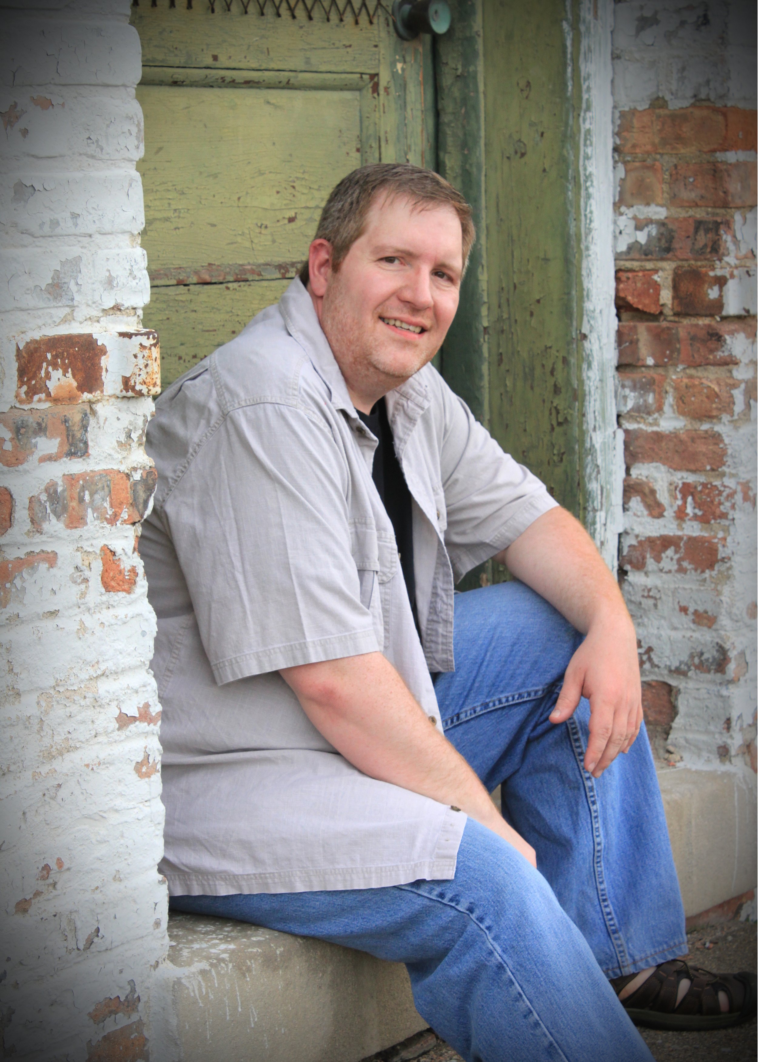 A man sitting on the ground with his back against a brick and weathered green wooden wall, smiling at the camera.
