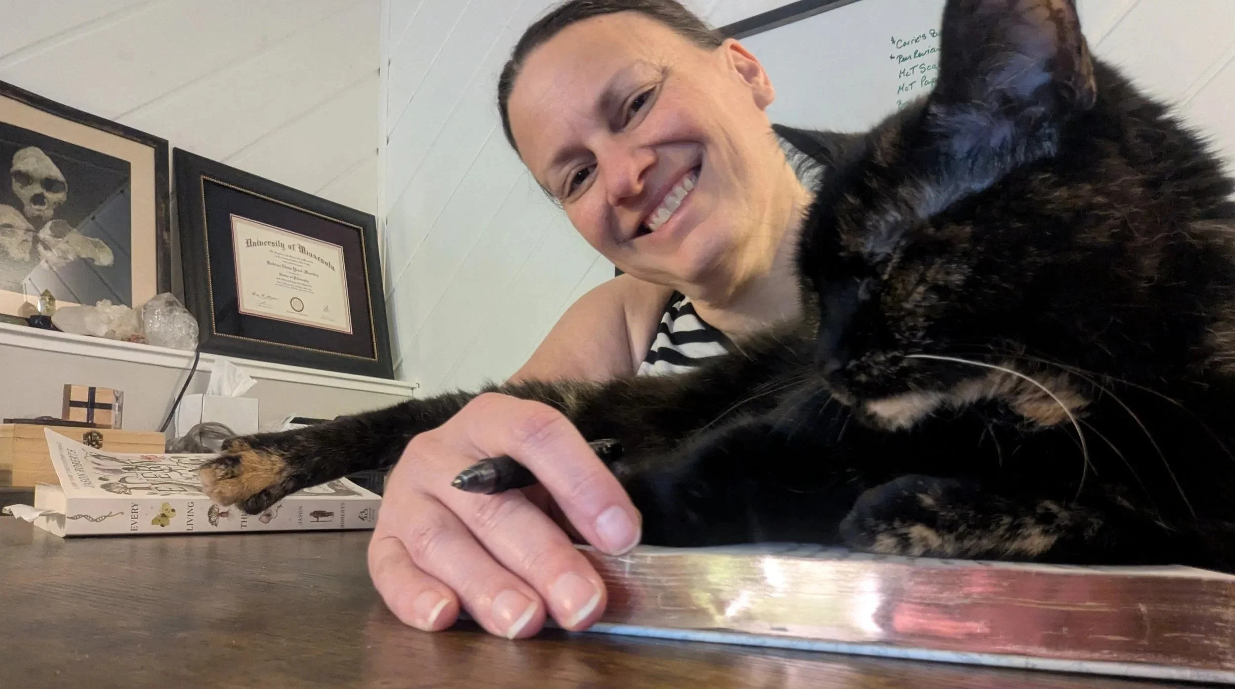 A woman smiles beside a black cat sitting on her open journal at a table, with books and writing materials nearby.