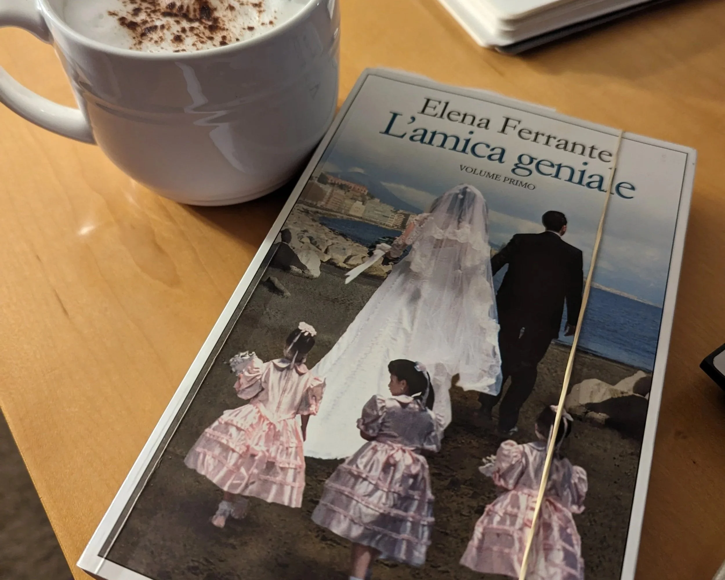 A cappuccino in a white mug sitting on a table next to a copy of Elena Ferrante's L'amica geniale.