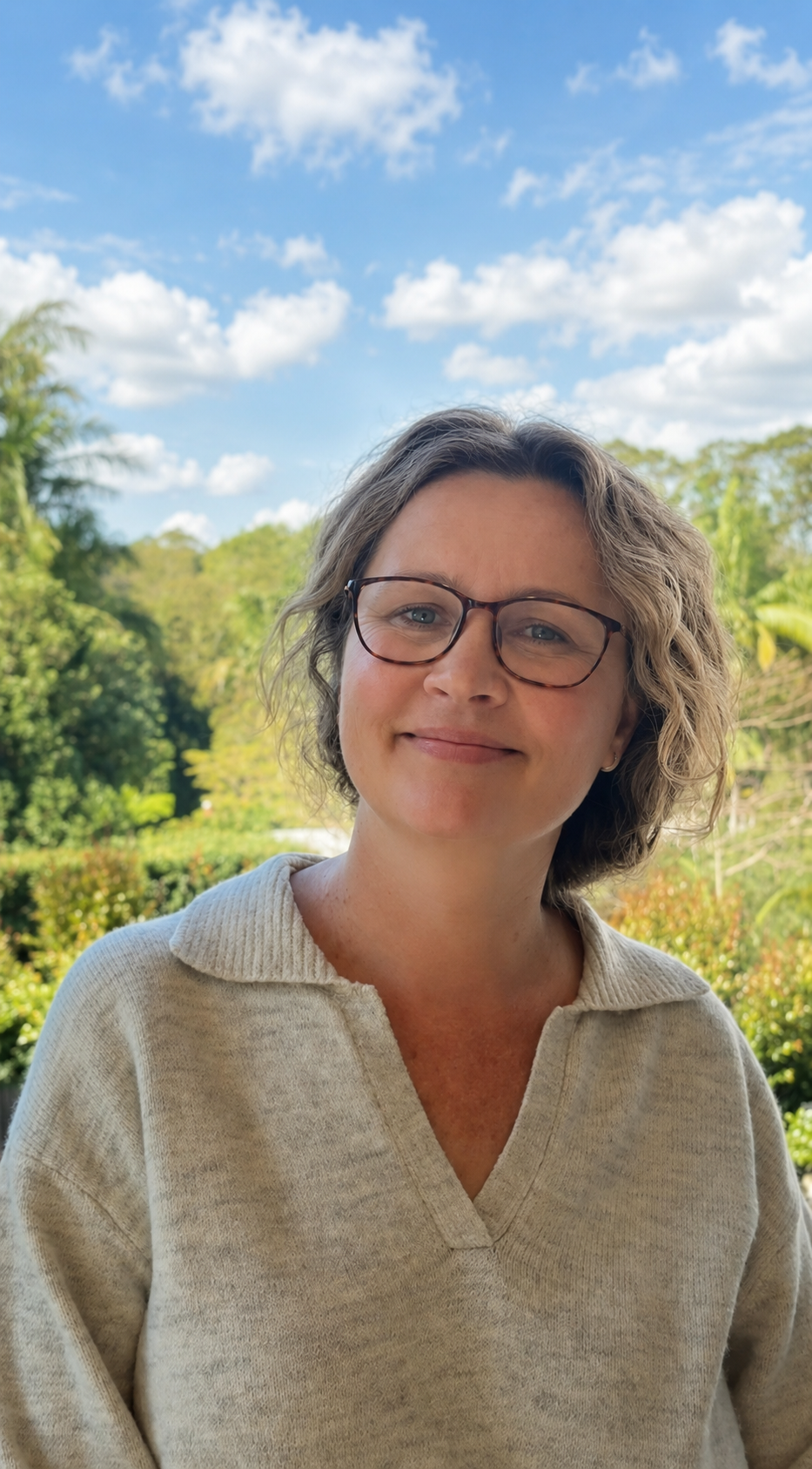 Portrait of a woman with short wavy hair and glasses, smiling outdoors with a background of trees and a blue sky with clouds.