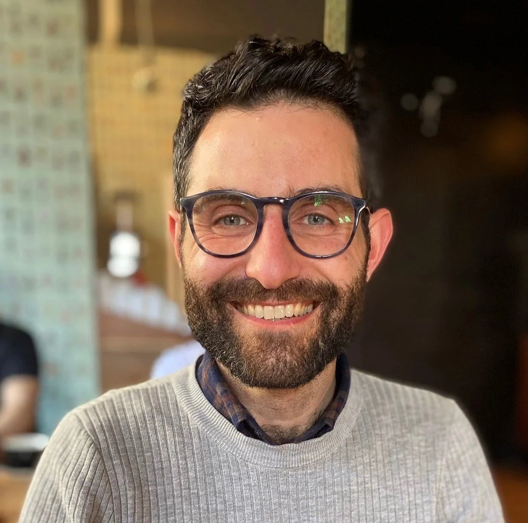A smiling man with glasses and a beard, wearing a light sweater over a collared shirt, in an indoor setting.