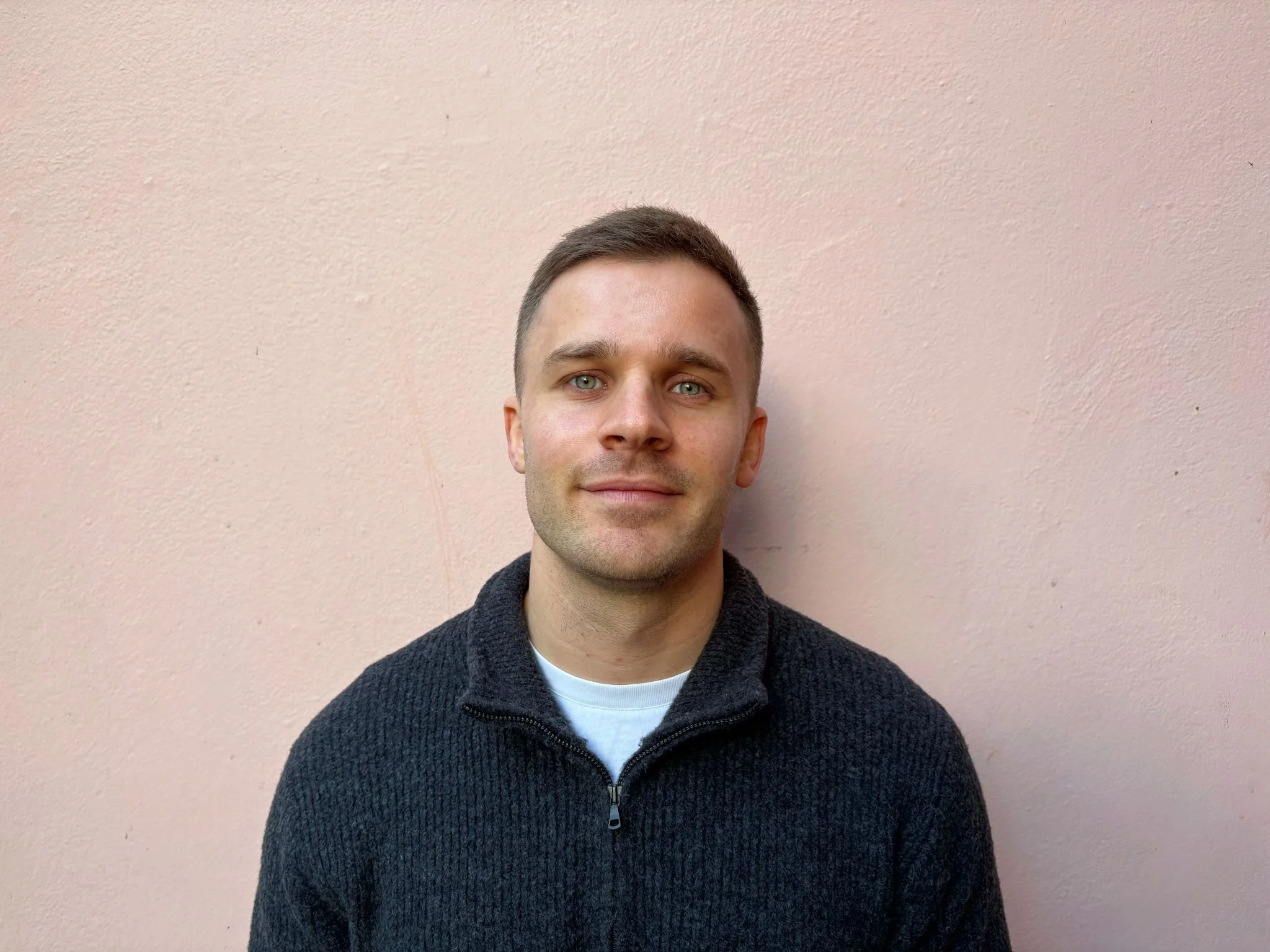 A portrait of a young man with short brown hair, blue eyes, and light skin, wearing a dark zip-up sweater over a white shirt, standing against a pale pink wall.
