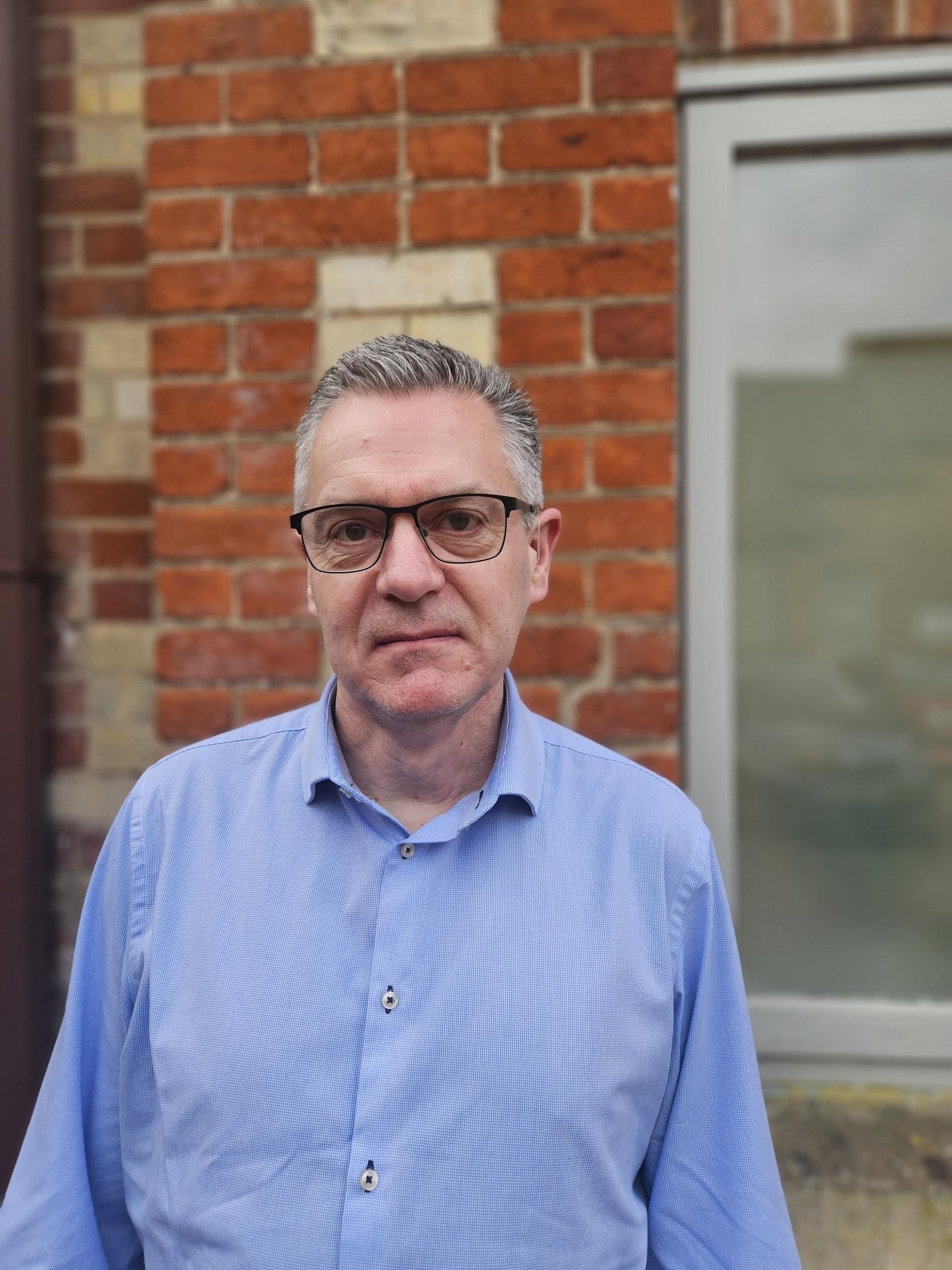 A middle-aged man with gray hair, glasses, and wearing a light blue collared shirt standing outdoors in front of a brick wall and window.