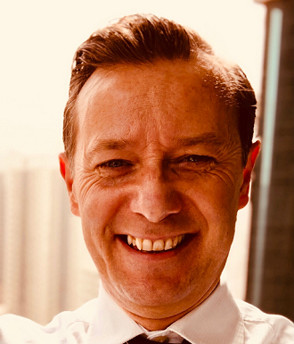 A smiling man with short, styled hair, wearing a white shirt outdoors with sunlight and a building in the background.