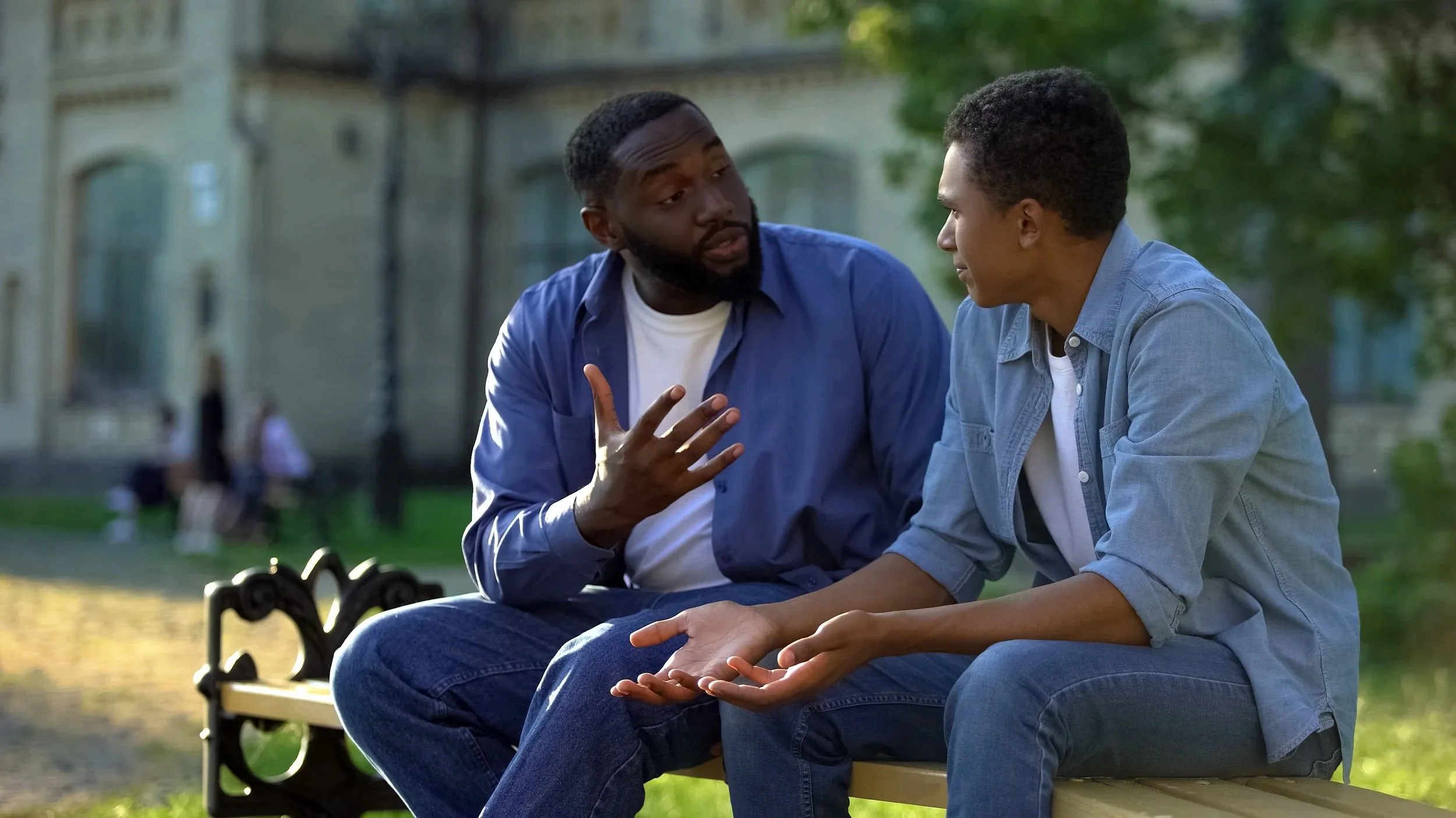 two men sitting on a bench talking to each other. Both are wearing white t-shirts with blue shirts