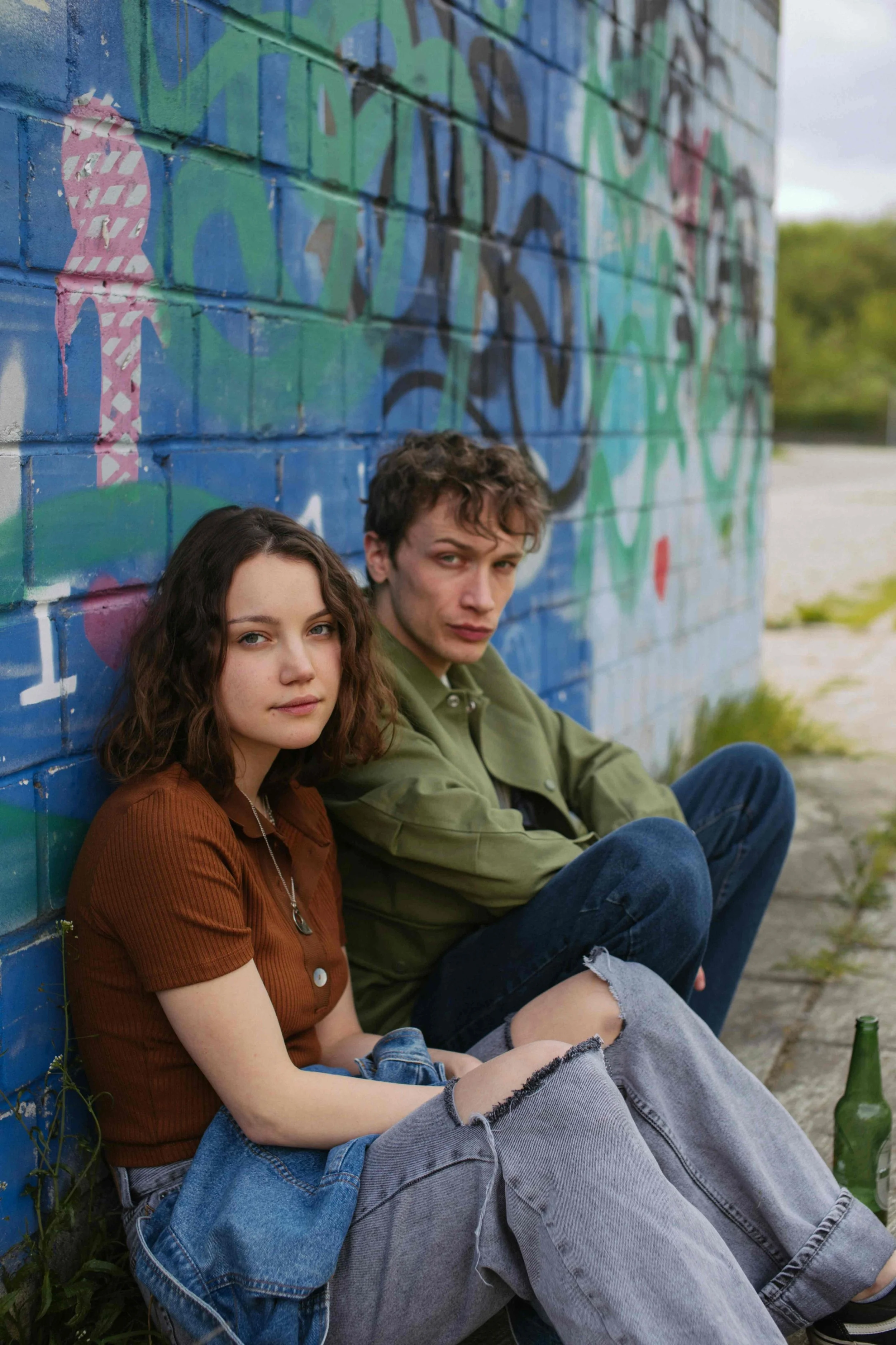 a young man and woman sitting on the gournd against a wall with colourful graffiti