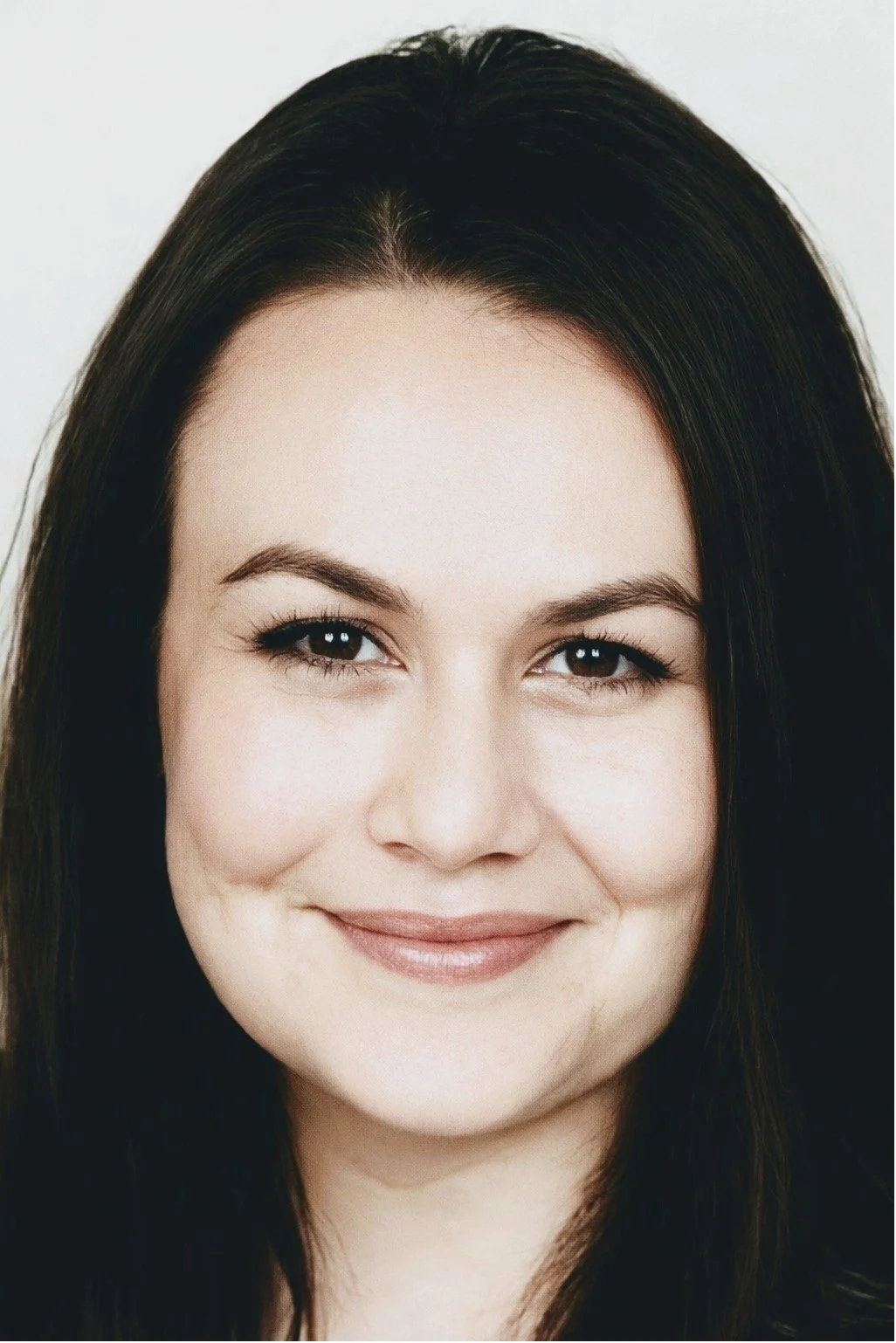 Close-up portrait of a smiling woman with dark brown hair, light skin, and light makeup against a plain background.
