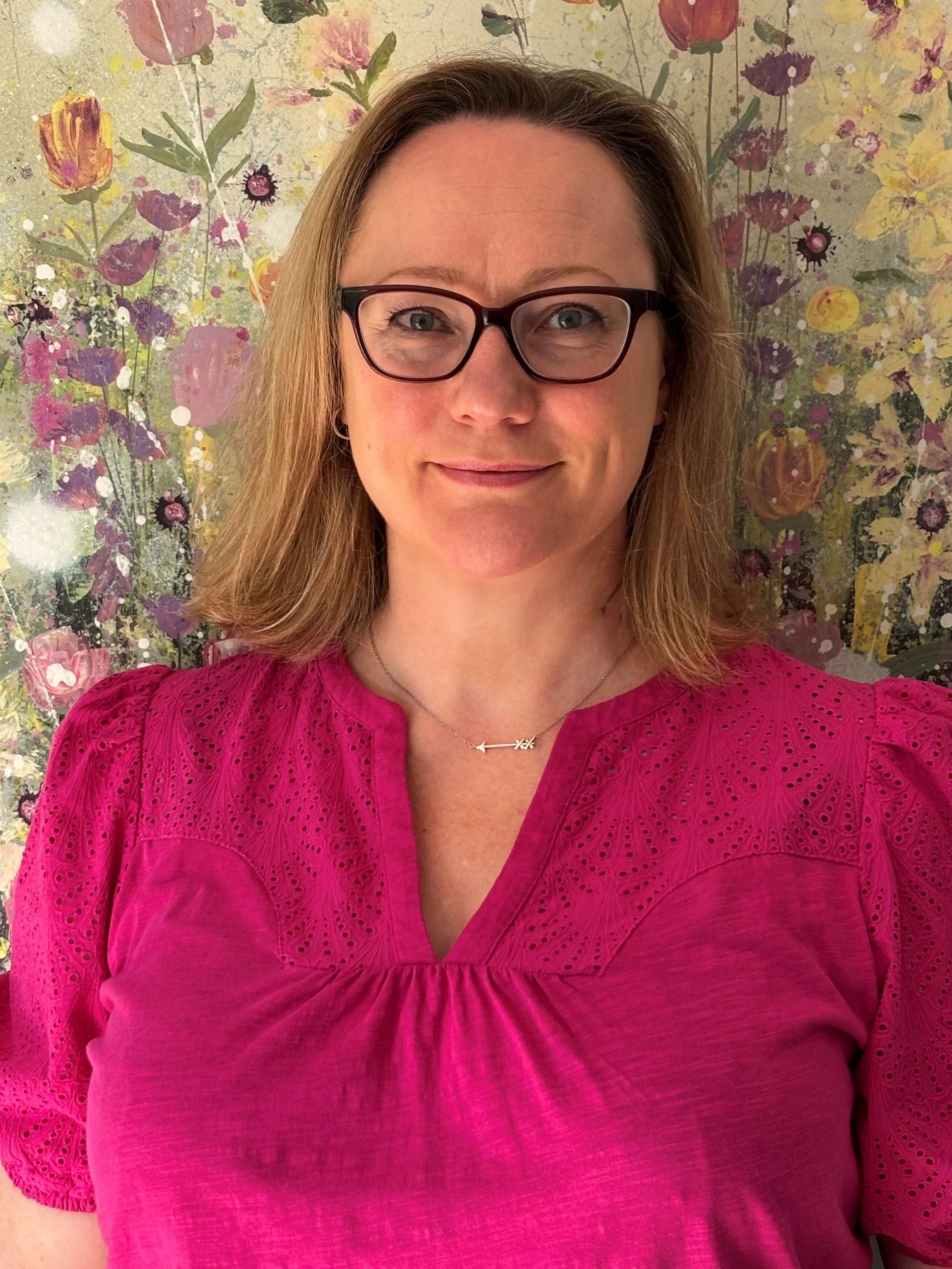 A woman with shoulder-length blonde hair, glasses, and a pink embroidered blouse stands in front of a floral watercolor painting with pink, purple, yellow, and green colors.