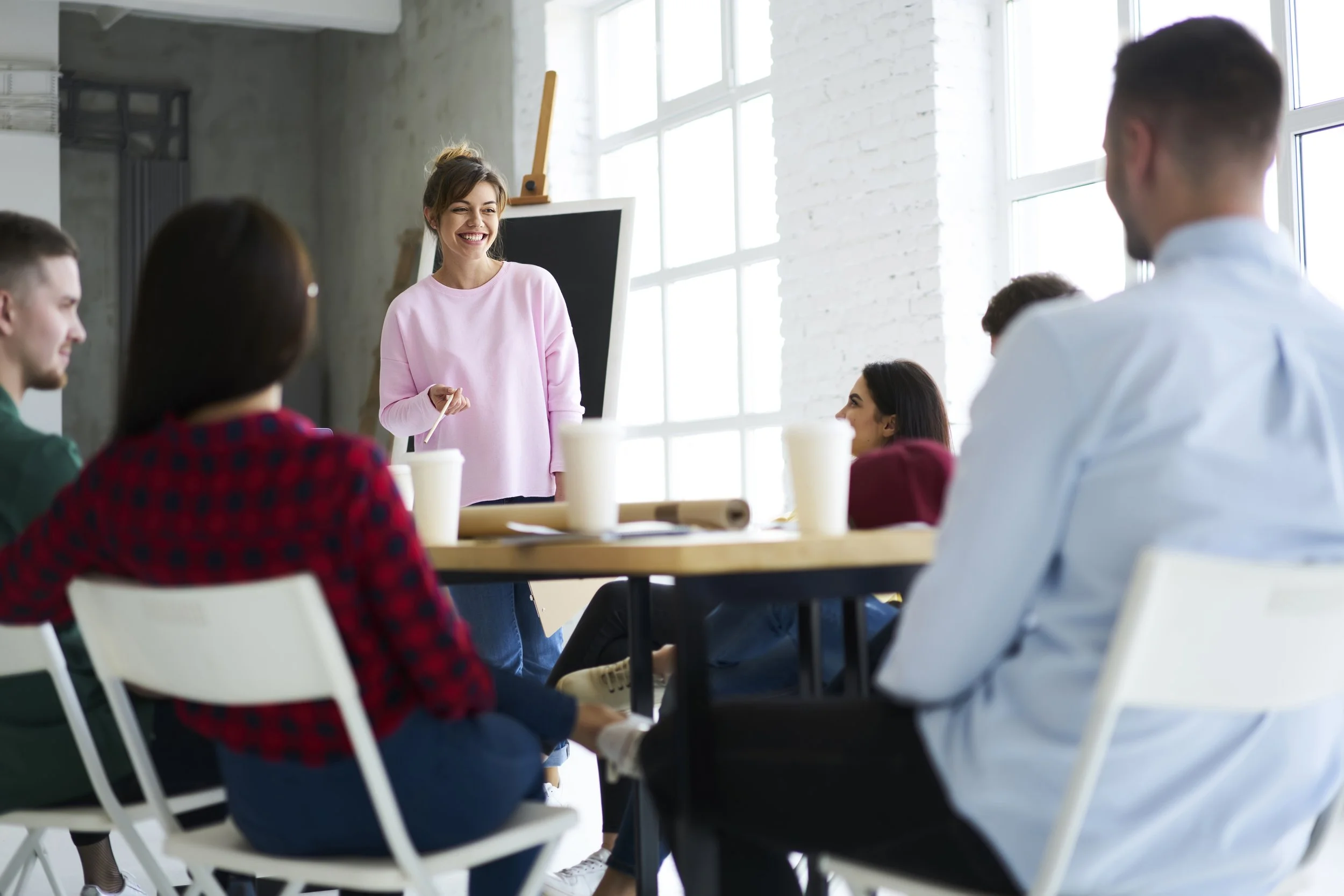 six people in a meeting around a table. A woman in a pink sweater is standing presenting