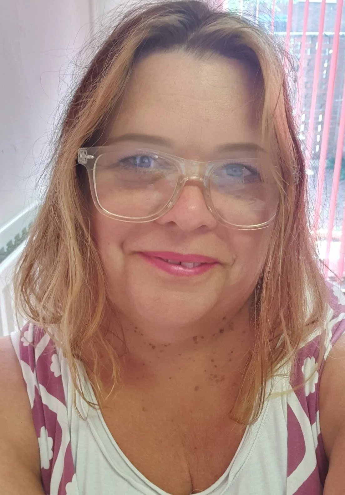 A woman with shoulder-length light brown hair, wearing large clear glasses, pink lipstick, and a white tank top with pink floral design. She is smiling indoors with pink blinds and a window in the background.