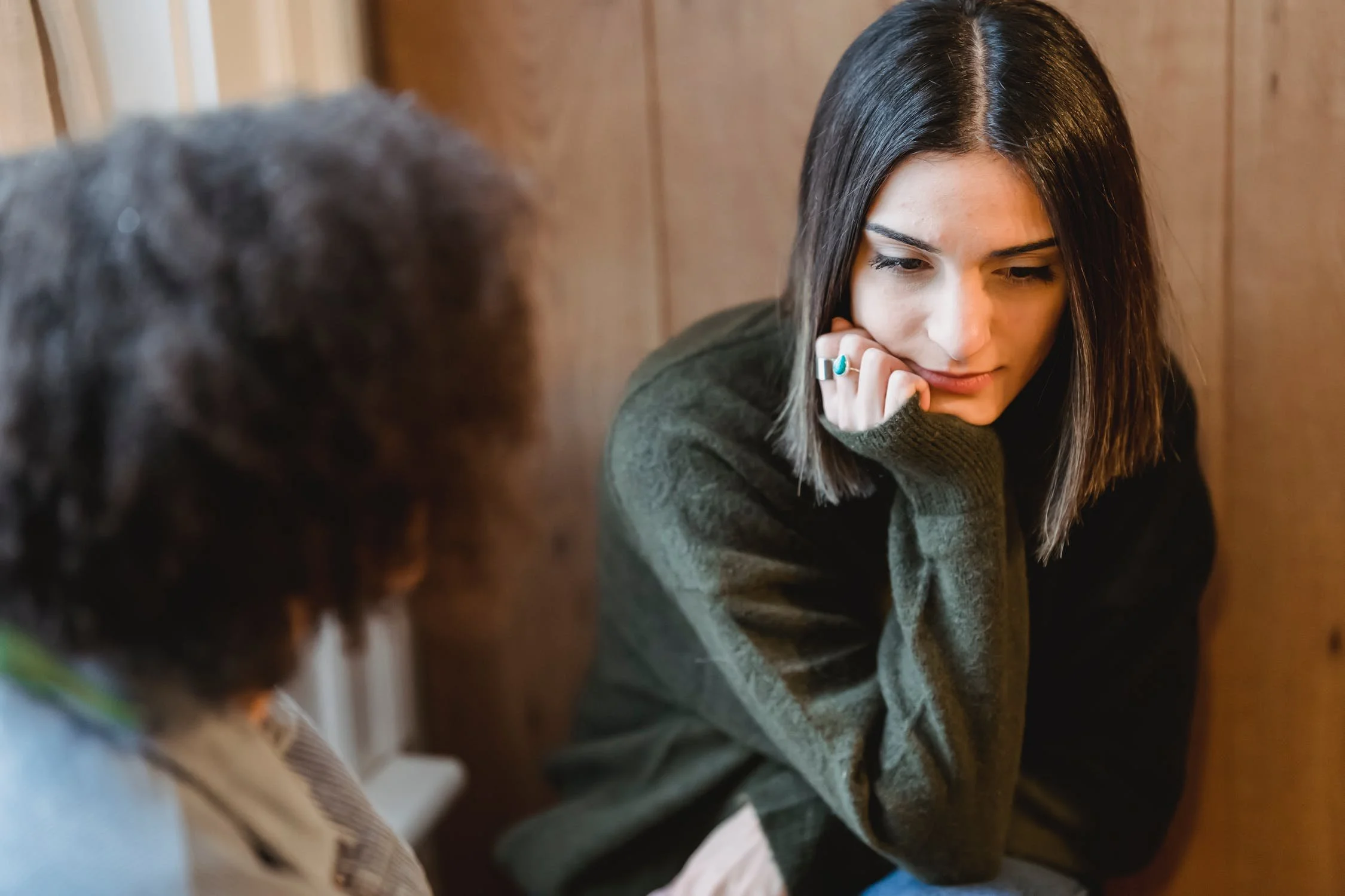 woman with straight brown hair sitting opposite another woman and looking at the ground