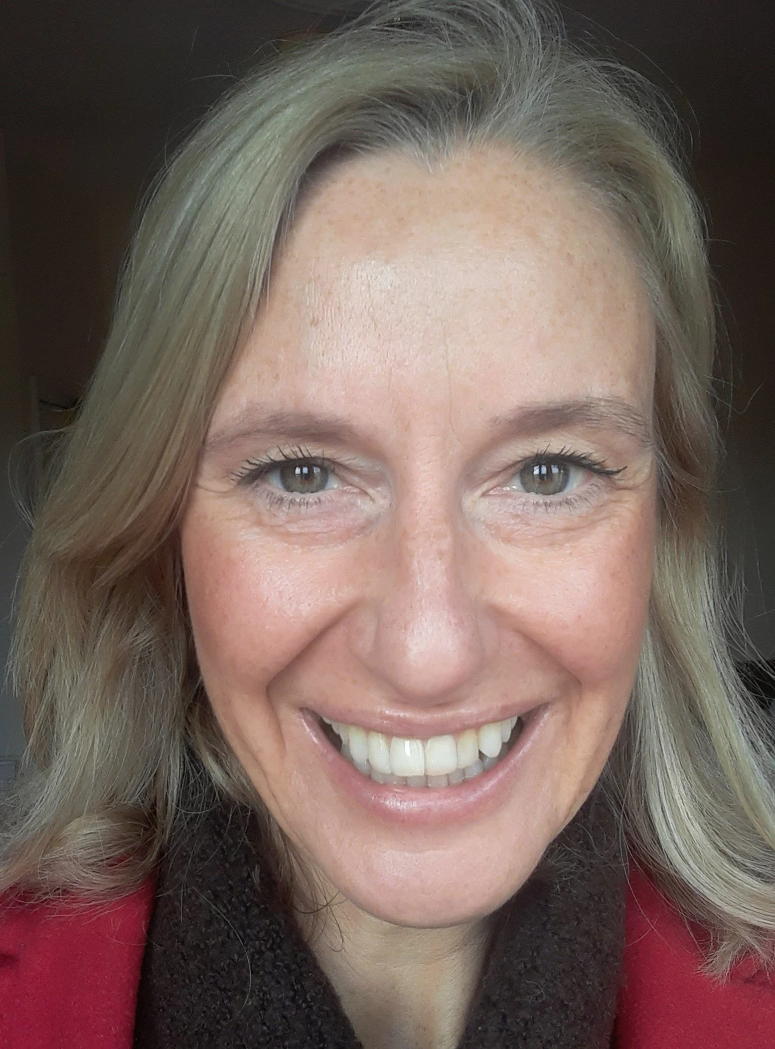 Close-up of smiling woman with blonde hair, light skin, blue eyes, wearing a red top and a dark scarf.