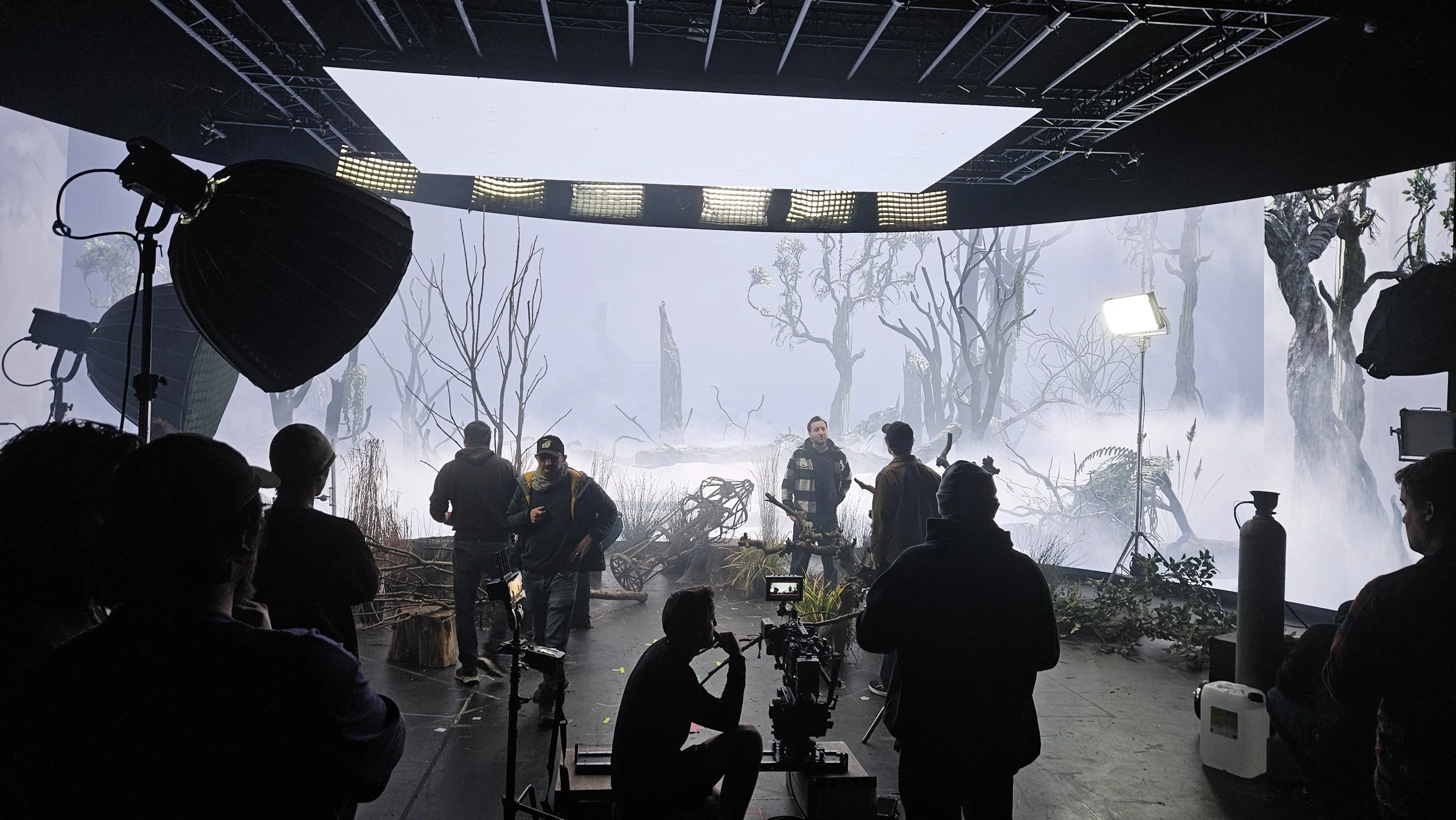 Film set with crew preparing for scene, featuring trees and fog background, professional lighting equipment, and large LED screens.
