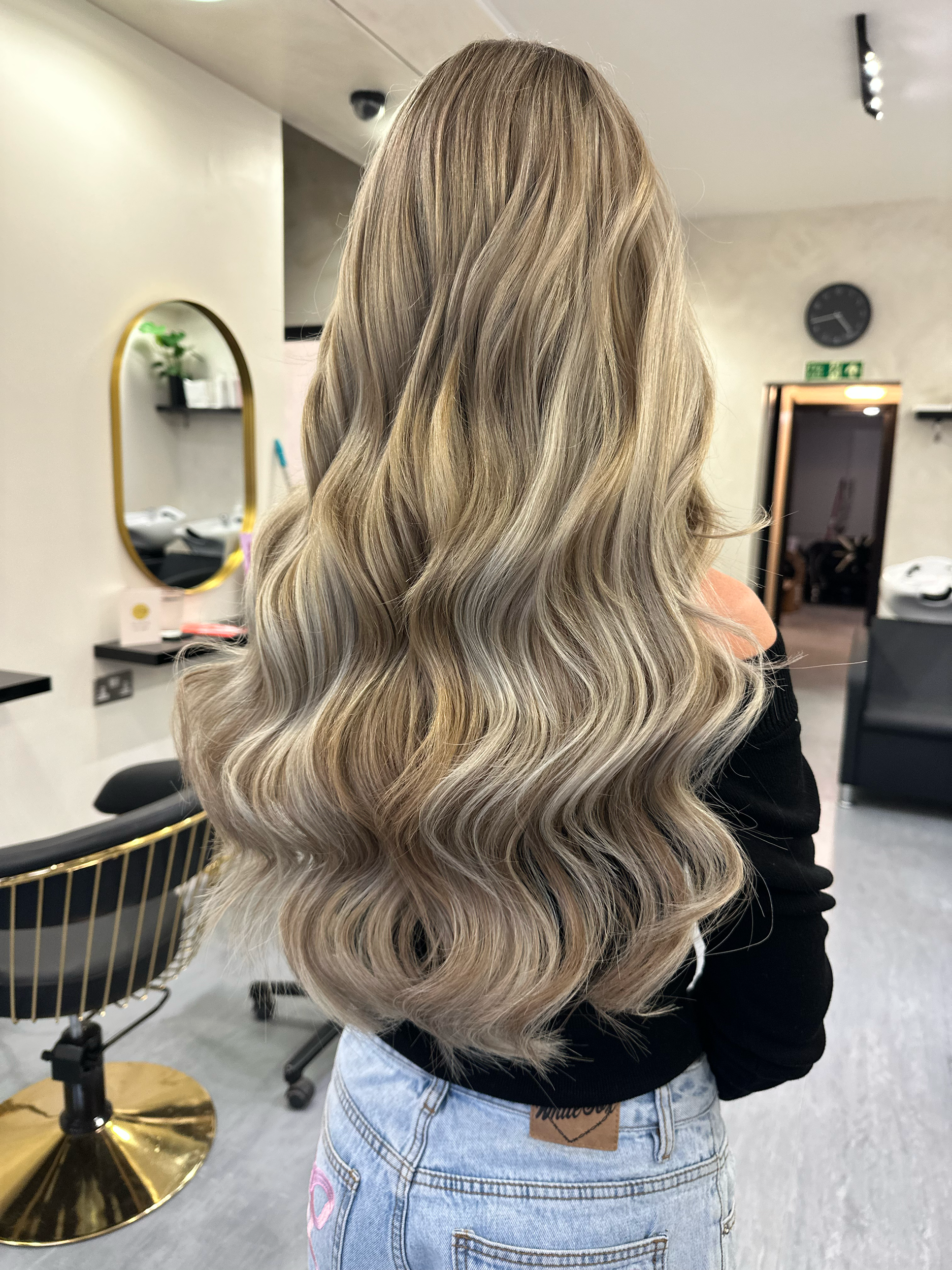 Back view of a woman with long, wavy, blonde hair in a salon.
