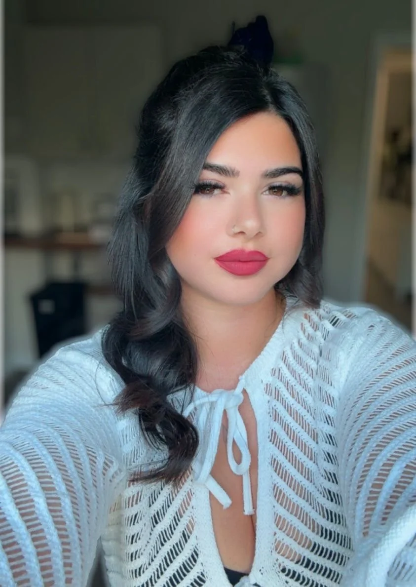 A woman with dark, wavy hair and makeup, wearing a white, see-through, striped top with a bow tie at the front, taking a selfie indoors.