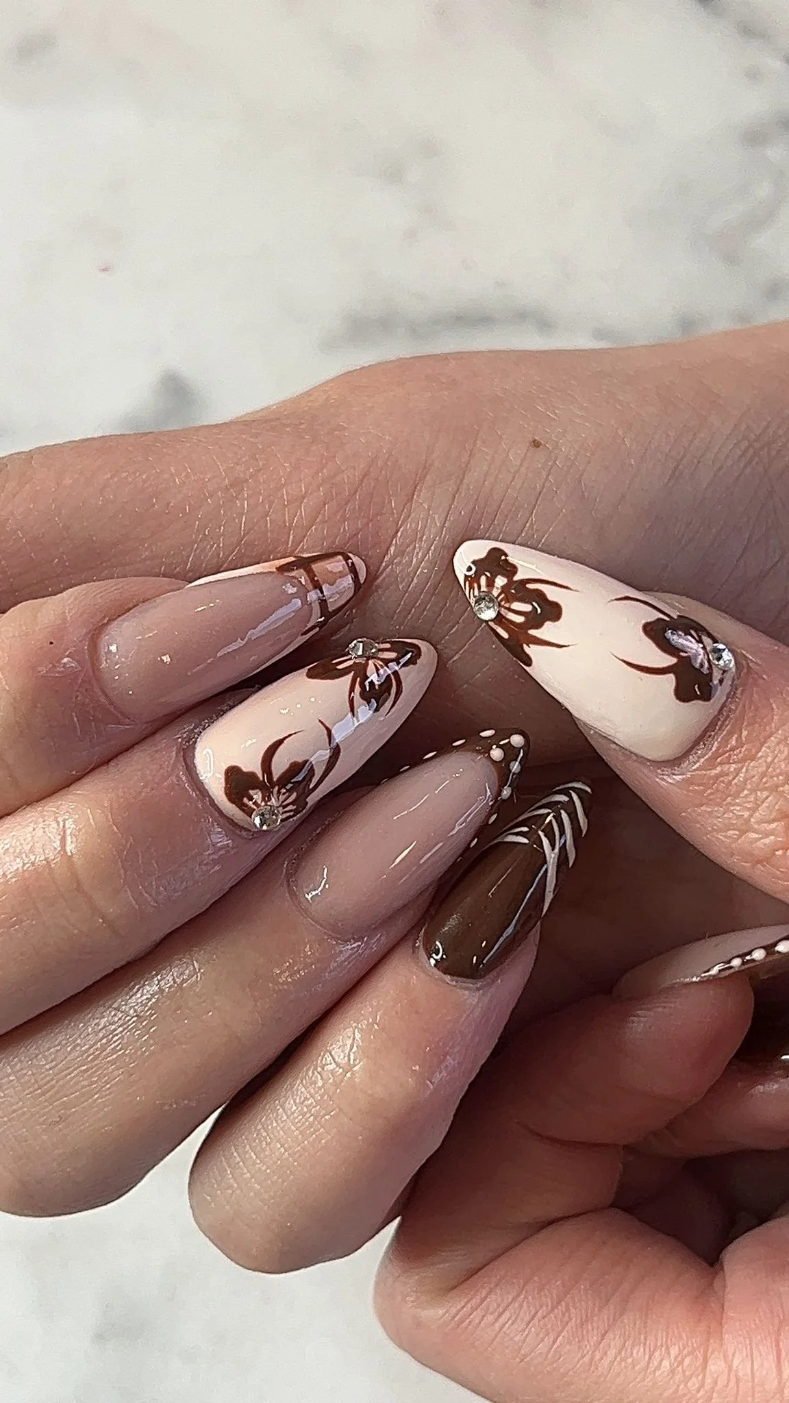 Close-up of manicured fingernails with a brown and white floral design, rhinestones, and striped patterns.