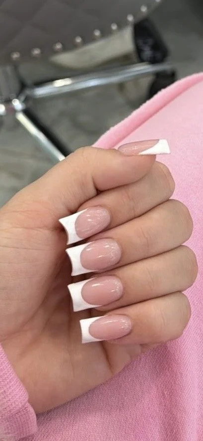 Close-up of a hand with manicured nails painted in light pink, held above a pink fabric surface, with a blurred metal chair in the background.