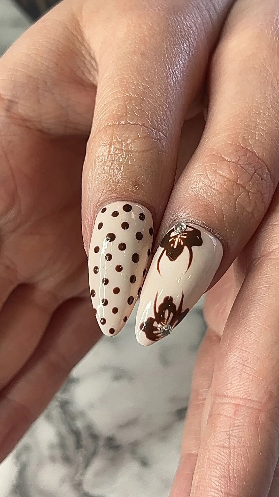 Close-up of a hand with two decorated nails: one with black polka dots on a nude background, the other with black spider gel designs and small rhinestones.