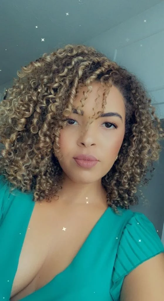 A woman with shoulder-length, curly blonde hair and light skin taking a selfie indoors. She is wearing a teal top with puffed sleeves and has a slight smile.