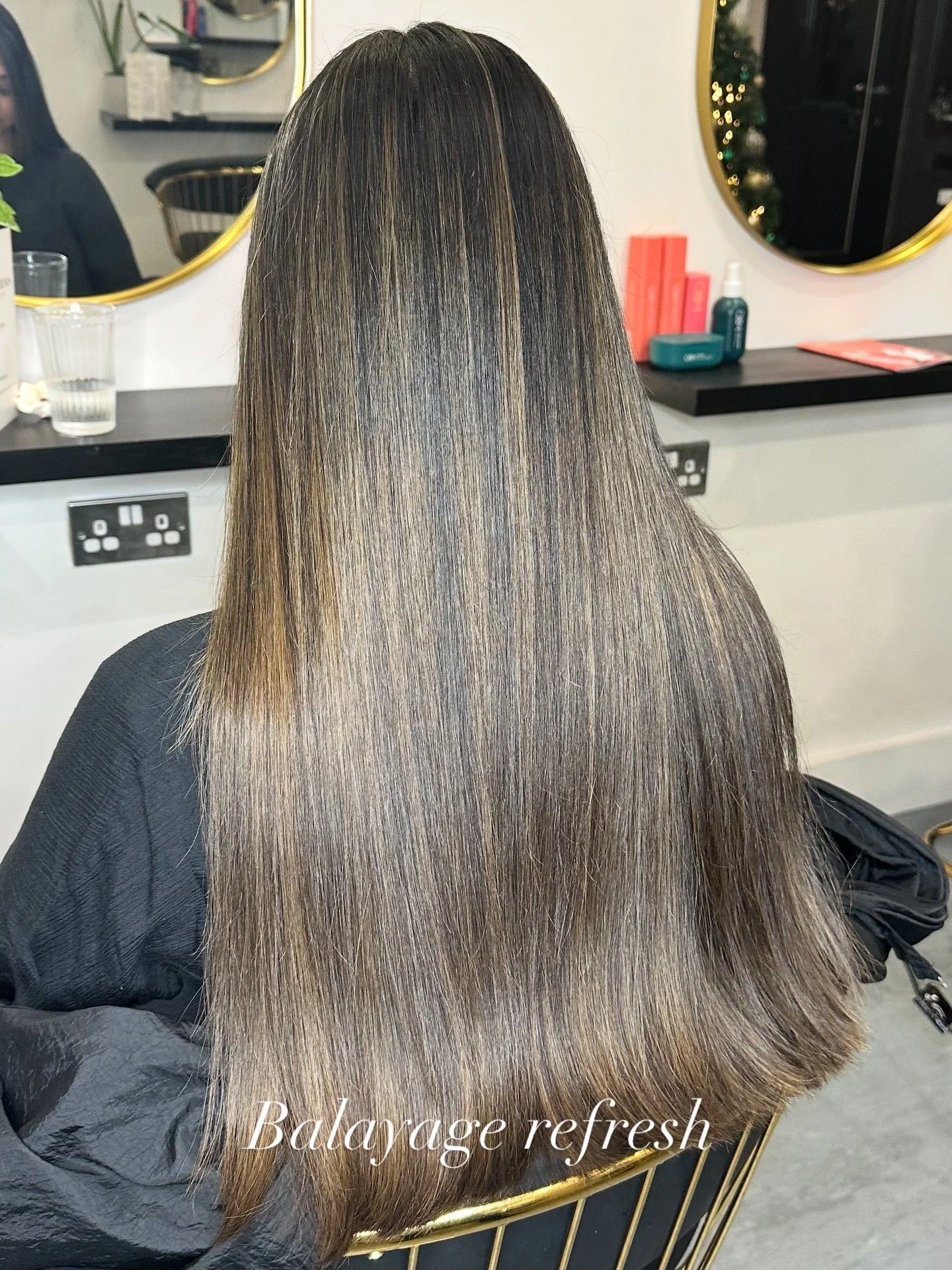 Back view of a woman with long, straight, shiny, and smooth hair that is freshly balanced and refreshed, sitting in a salon chair with a black cape around her shoulders, in front of a mirror and salon station.