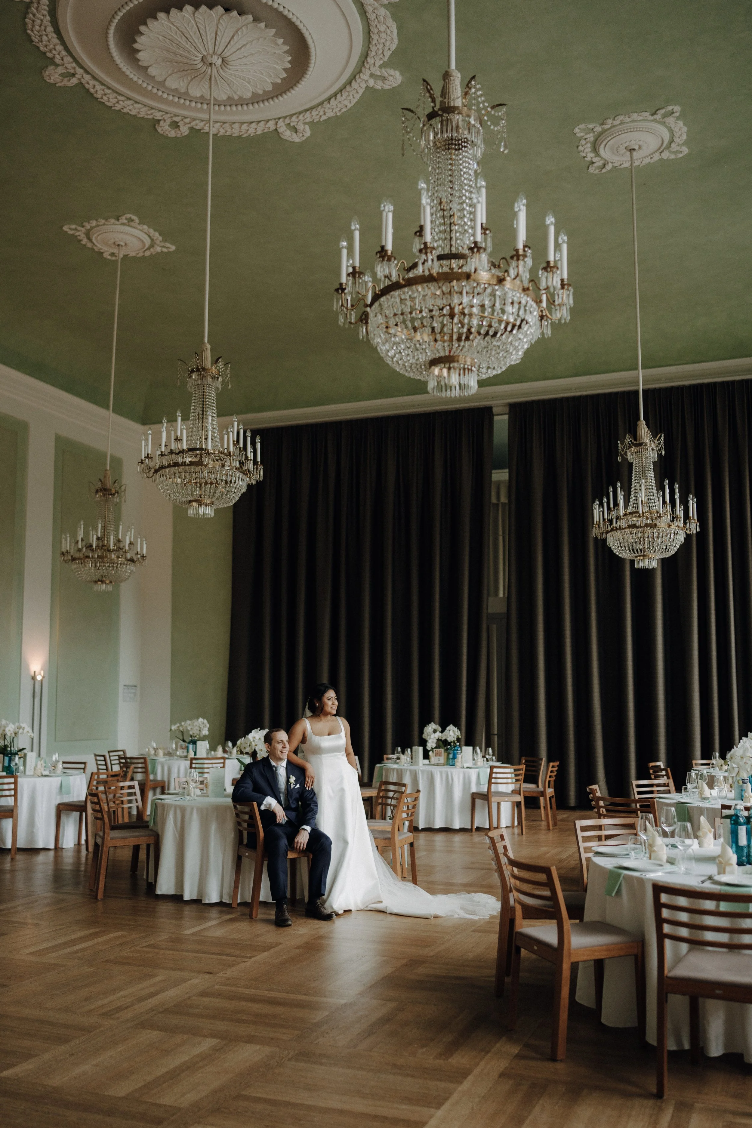 Ein Brautpaar posiert in einem eleganten, festlich gedeckten Saal mit Kronleuchtern und großen Vorhängen, während sie an ihrer Hochzeit feiern.