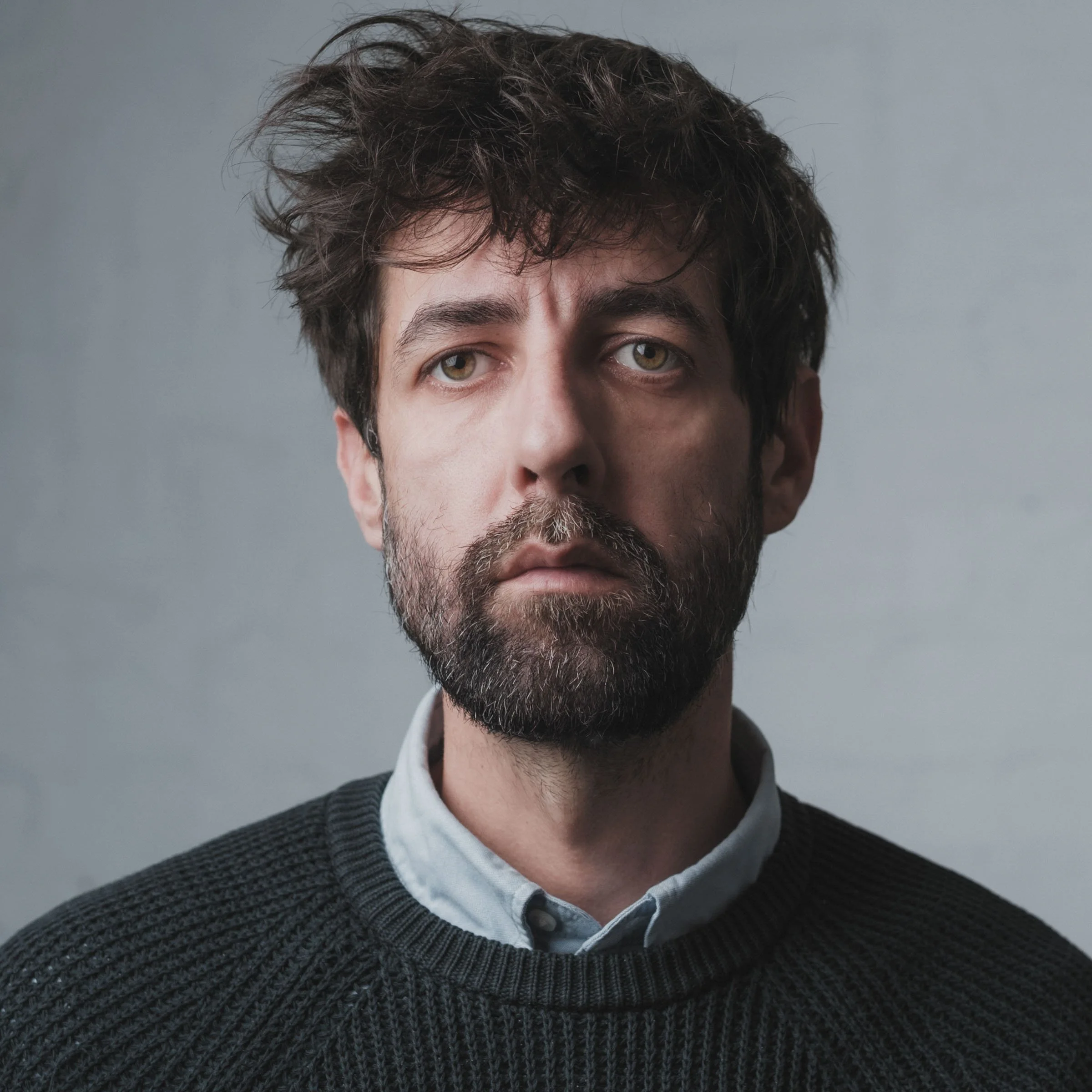 Un homme avec une expression neutre, portant un pull noir et une chemise blanche, face à un fond gris