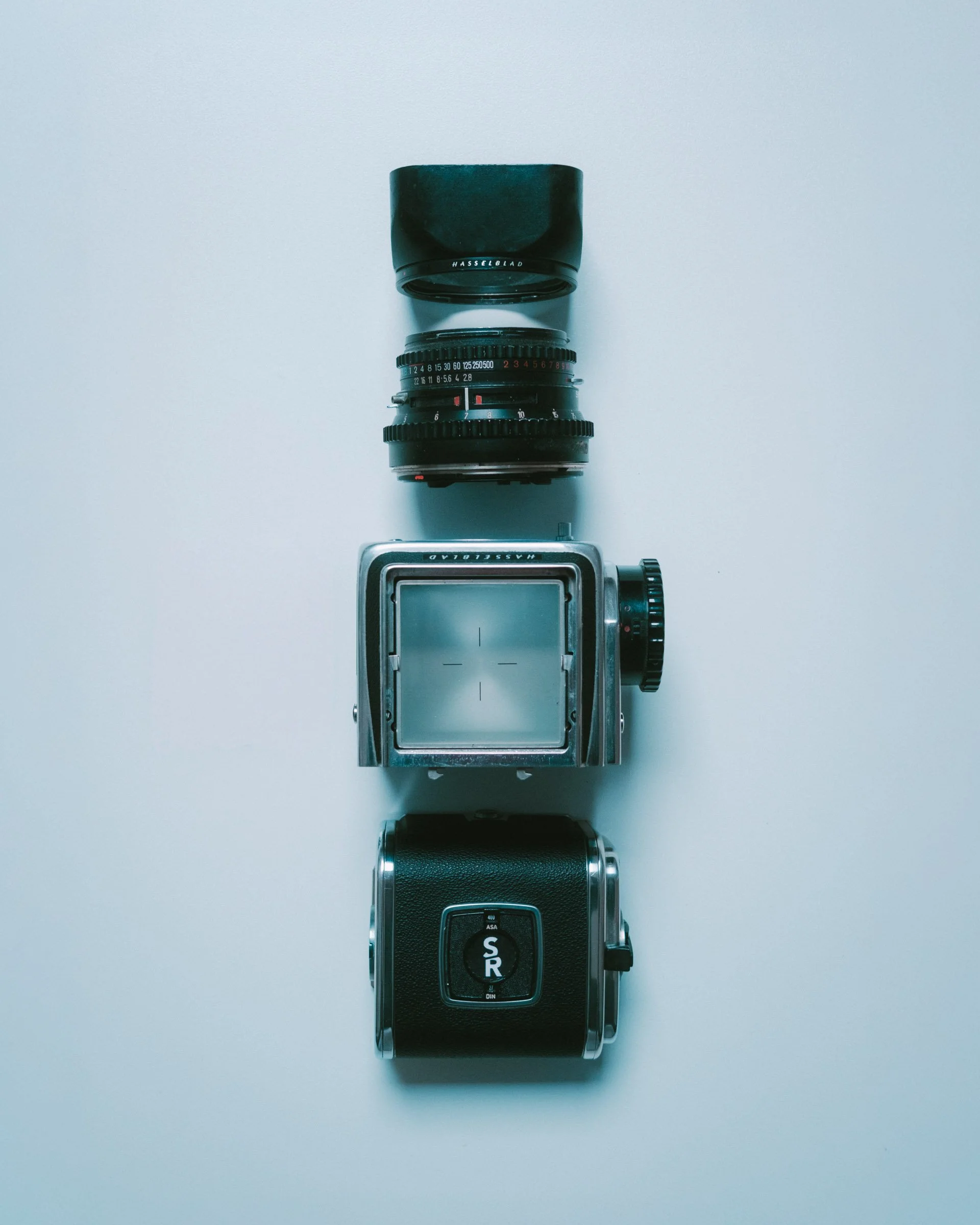 Une caméra Hasselblad démonté, un objectif, et un pare-soleil, tous alignés verticalement sur une surface blanche.