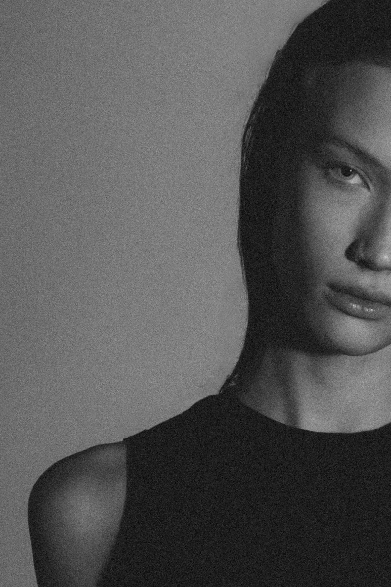 Portrait en noir et blanc d'une femme avec une expression neutre, moitié du visage visible, cheveux mouillés ou lisses, portant un haut sans manches, fond simple